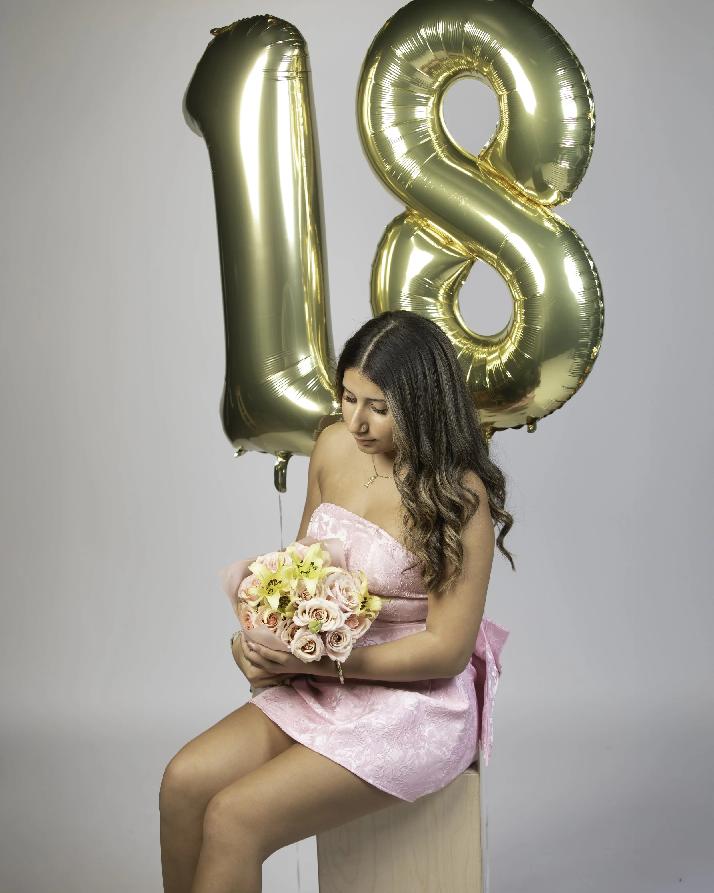 Young woman in pink dress holding a bouquet of flowers sitting in front of large gold '18' balloons opposed to a plain background.