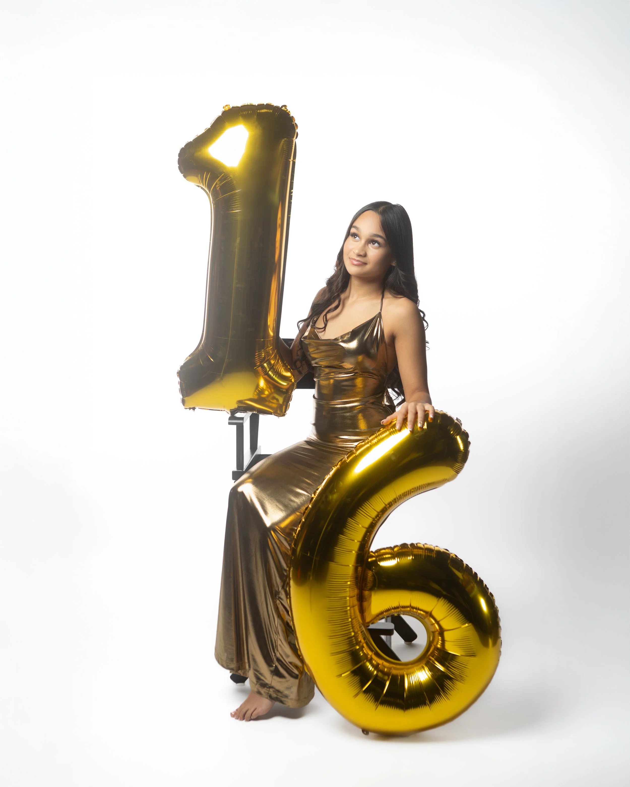 A woman in a shiny gold dress holding a large black and gold balloon in the shape of the number 1 and sitting behind a big gold balloon in the shape of the number 6, celebrating a 16th birthday.