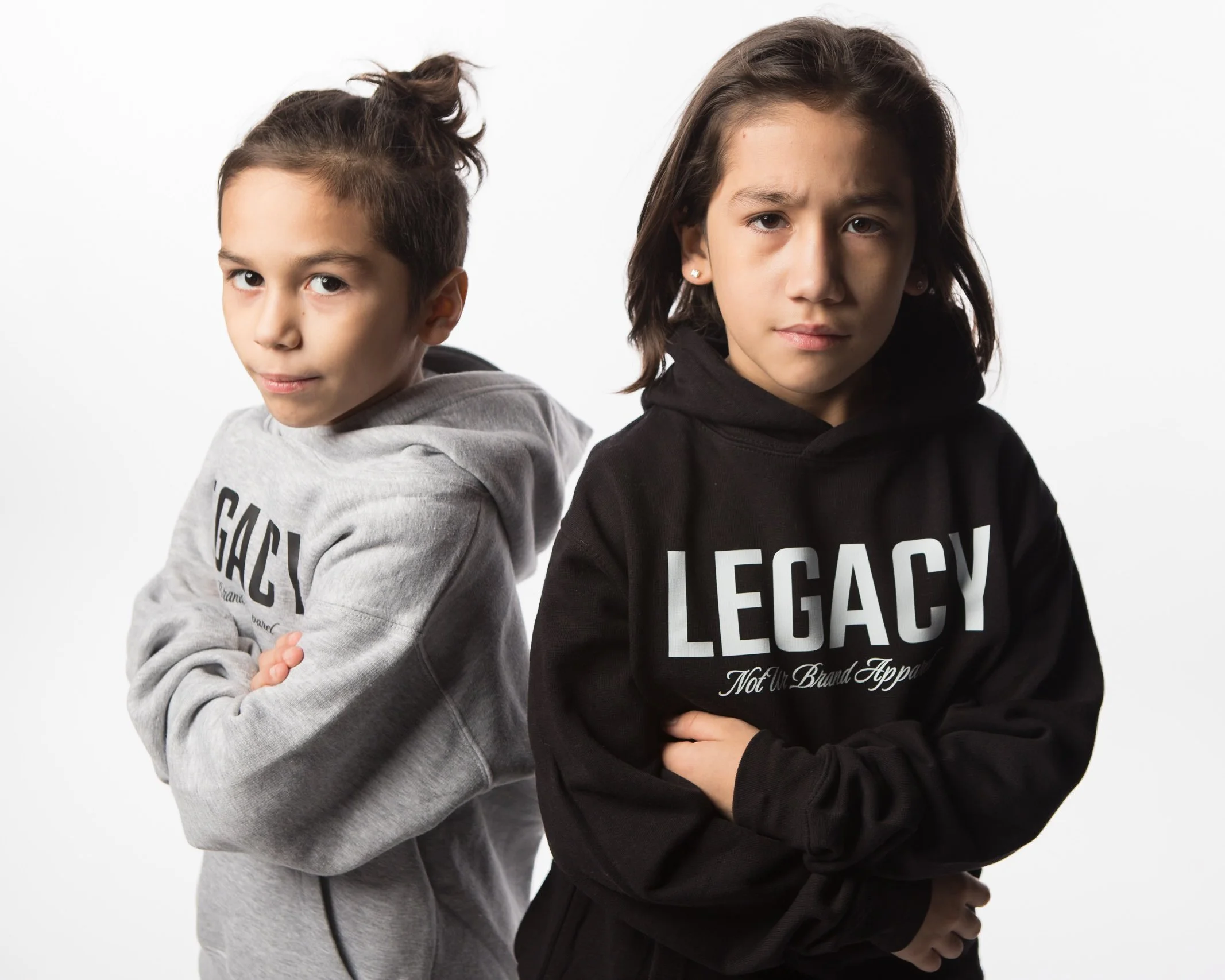 Two children, a boy and a girl, stand back to back with arms crossed, wearing hoodies with the word 'LEGACY' on them, in front of a white background.