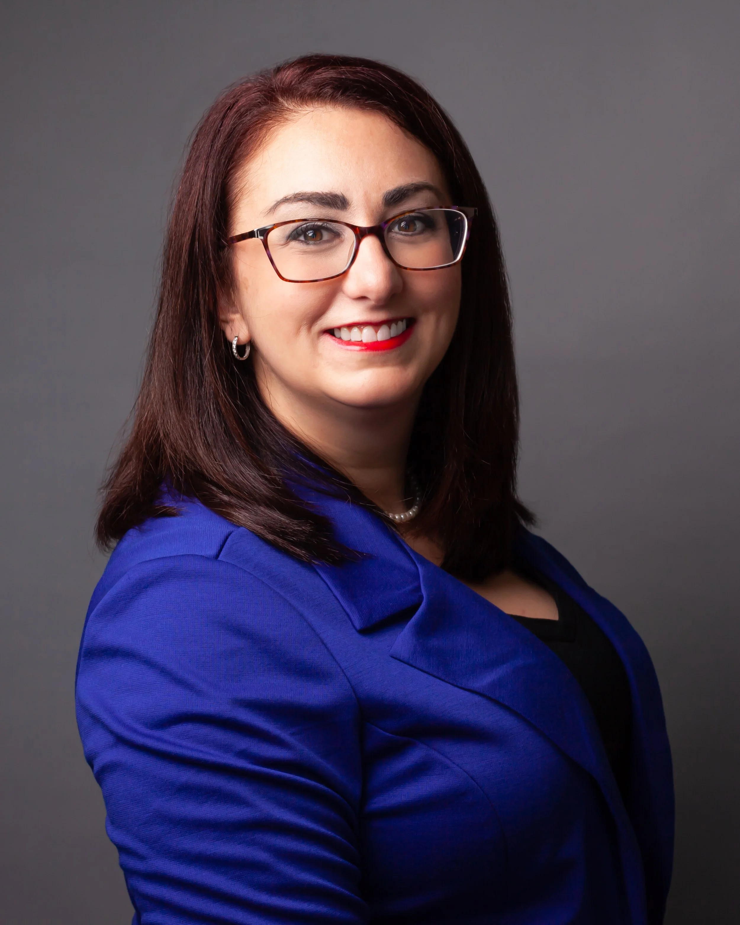 Professional woman with shoulder-length dark hair, glasses, pearl earrings, and red lipstick, wearing a royal blue blazer and smiling, against a gray background.