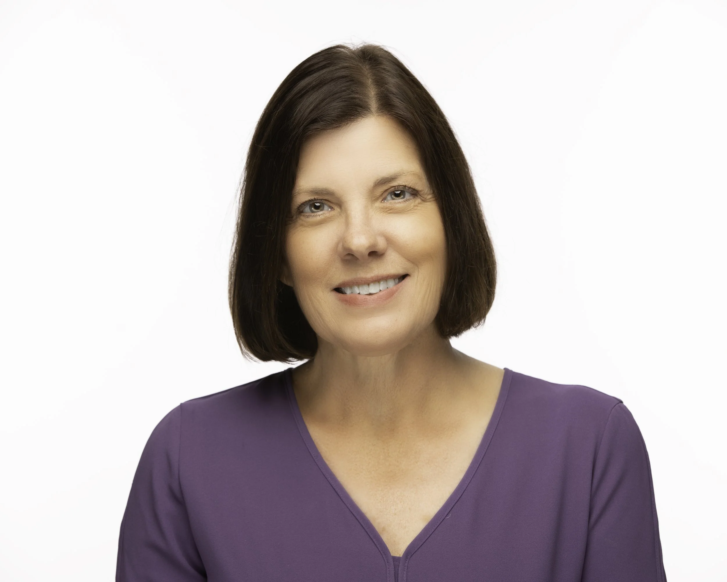 Portrait of a middle-aged woman with shoulder-length brown hair, smiling, wearing a purple blouse, against a plain white background.