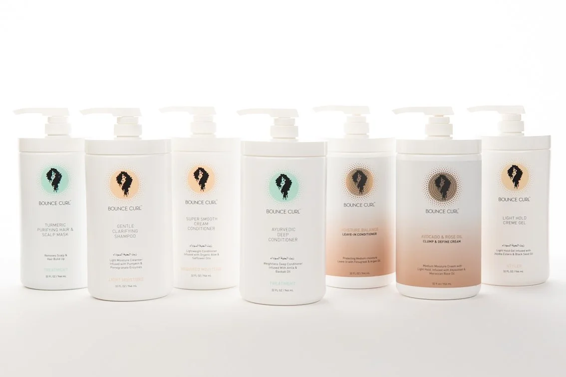 Seven white bottles of haircare products with pump dispensers from the Bounce Curl brand, labeled with different hair treatment types.