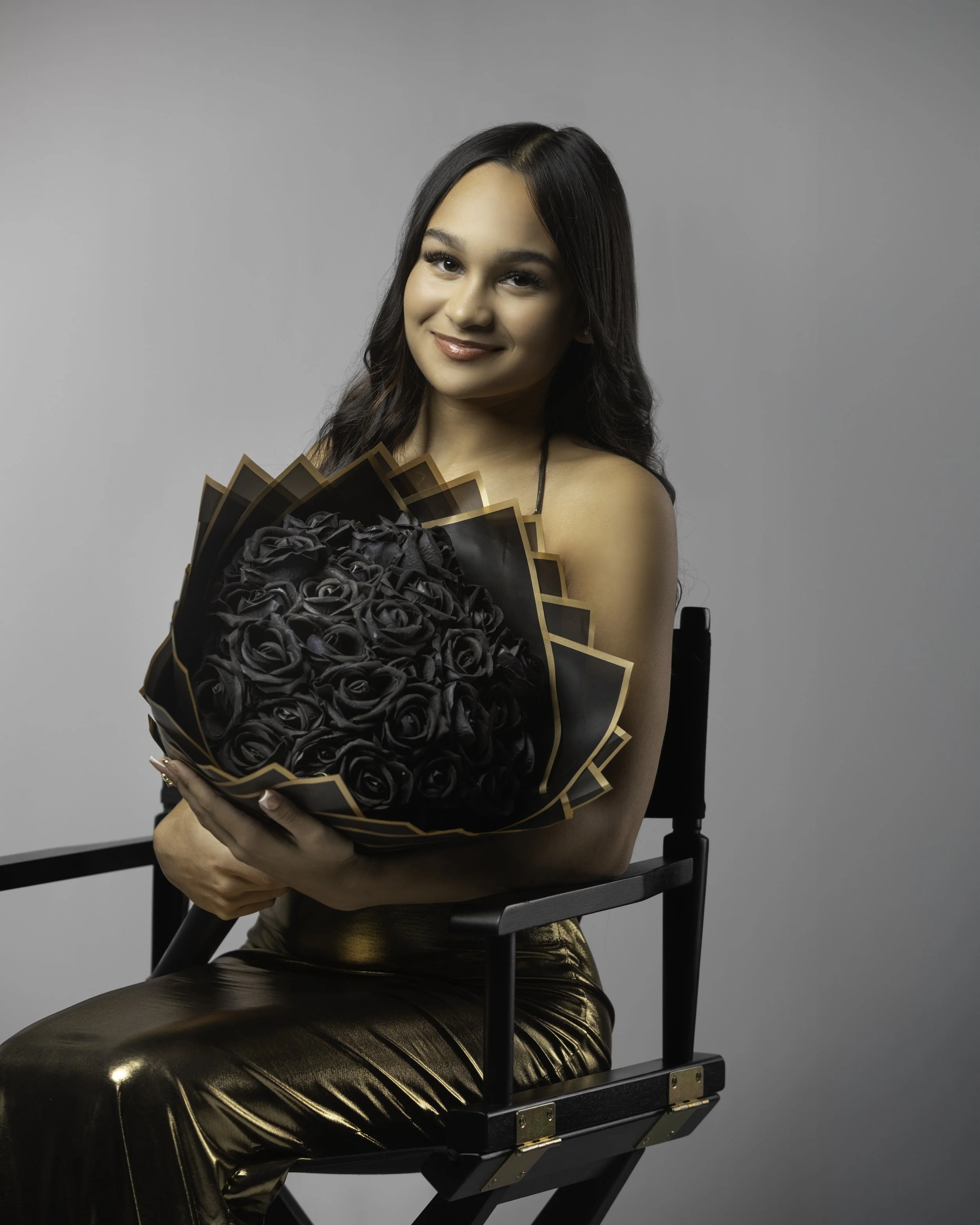 A woman sitting on a black director's chair, holding a bouquet of black roses wrapped in black and gold paper, against a plain light gray background, smiling at the camera.