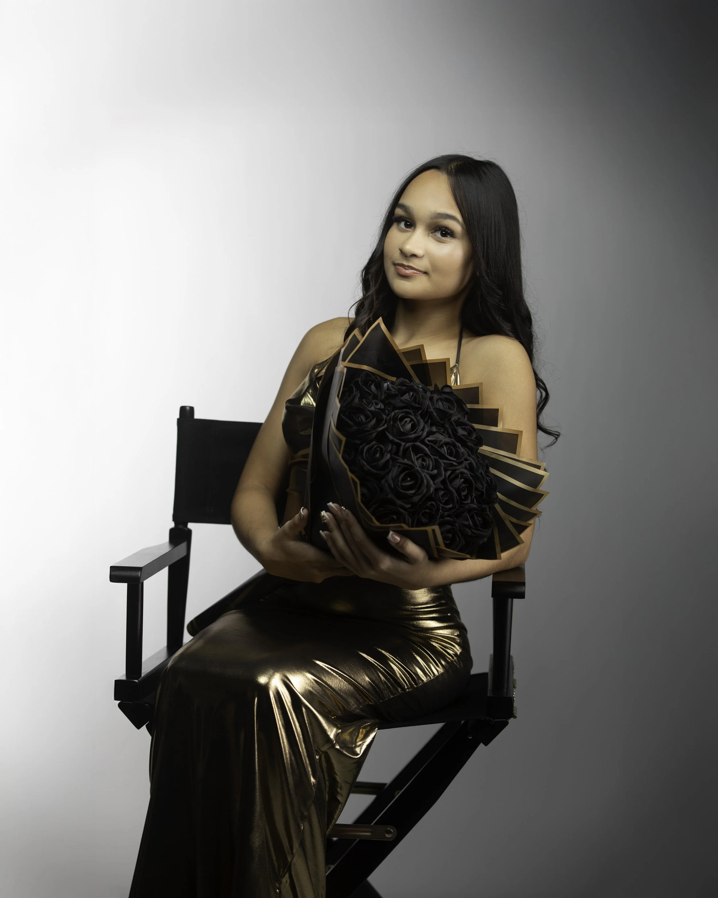 Young woman in a metallic gold dress sitting on a director's chair, holding a large bouquet of black roses with gold accents, against a neutral background.