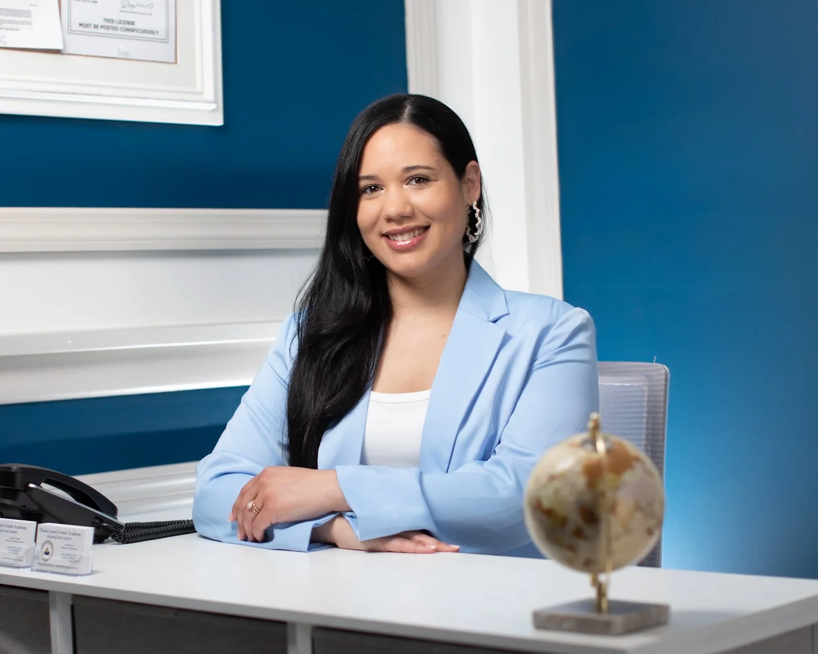 A woman with long black hair wearing a light blue blazer and earrings, sitting at a desk in an office with blue walls. There is a black telephone, business cards, and a small globe on the desk.