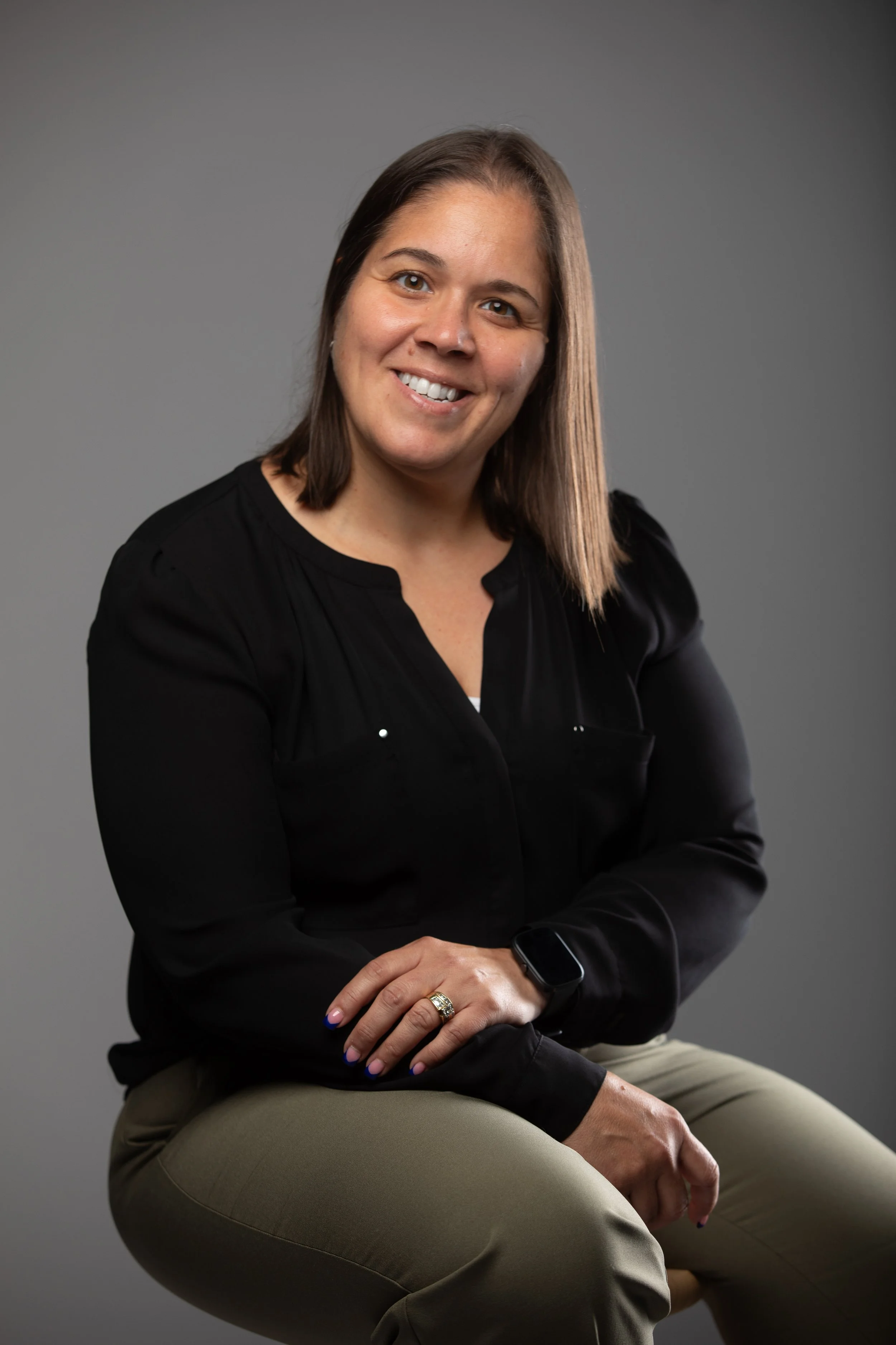 A woman with shoulder-length brown hair, smiling, wearing a black blouse and beige pants, sitting against a plain gray background.