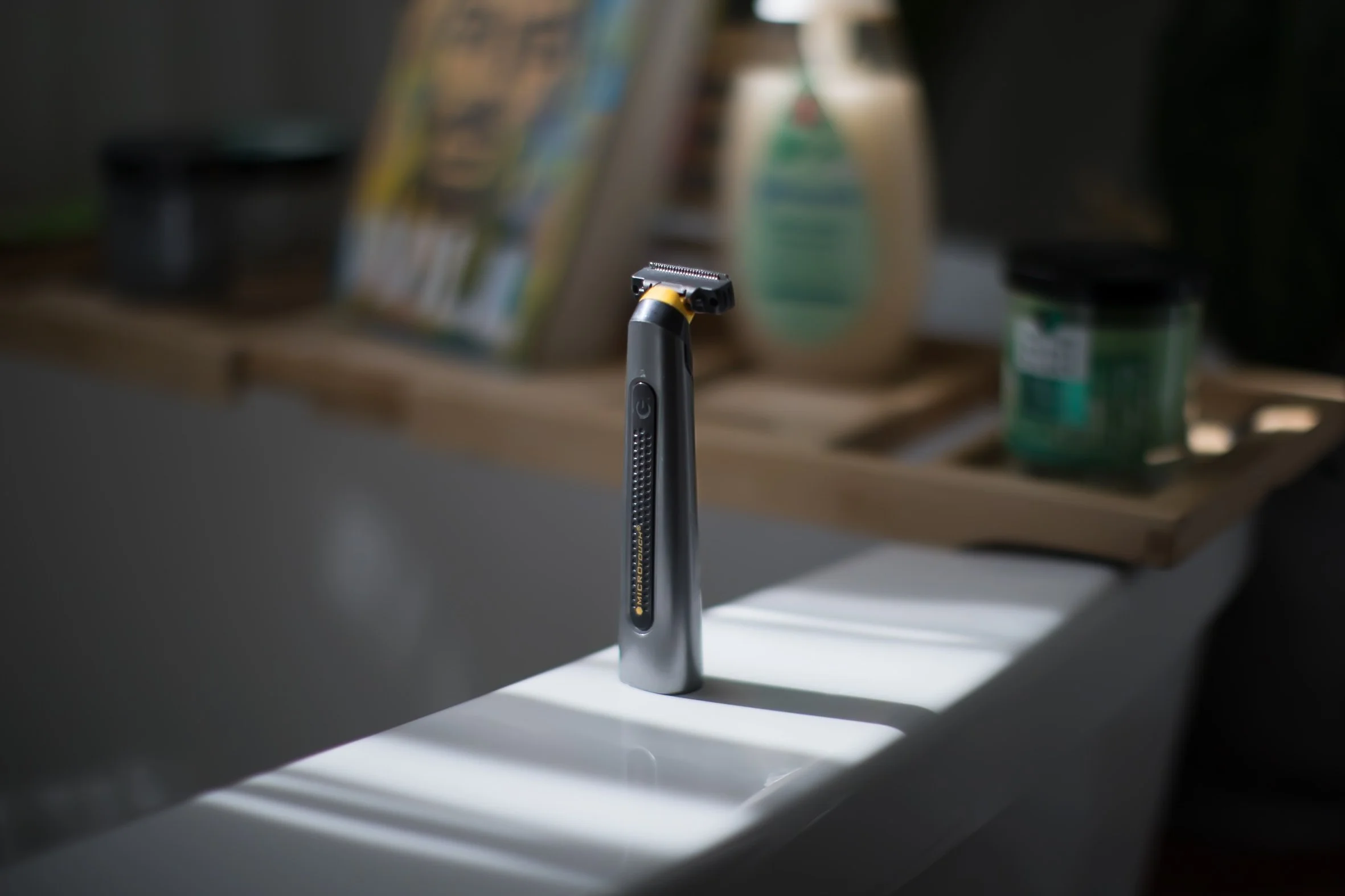 An electric razor standing upright on a white surface with shadows cast across it, with a blurred background showing a wooden tray with bottles and containers.
