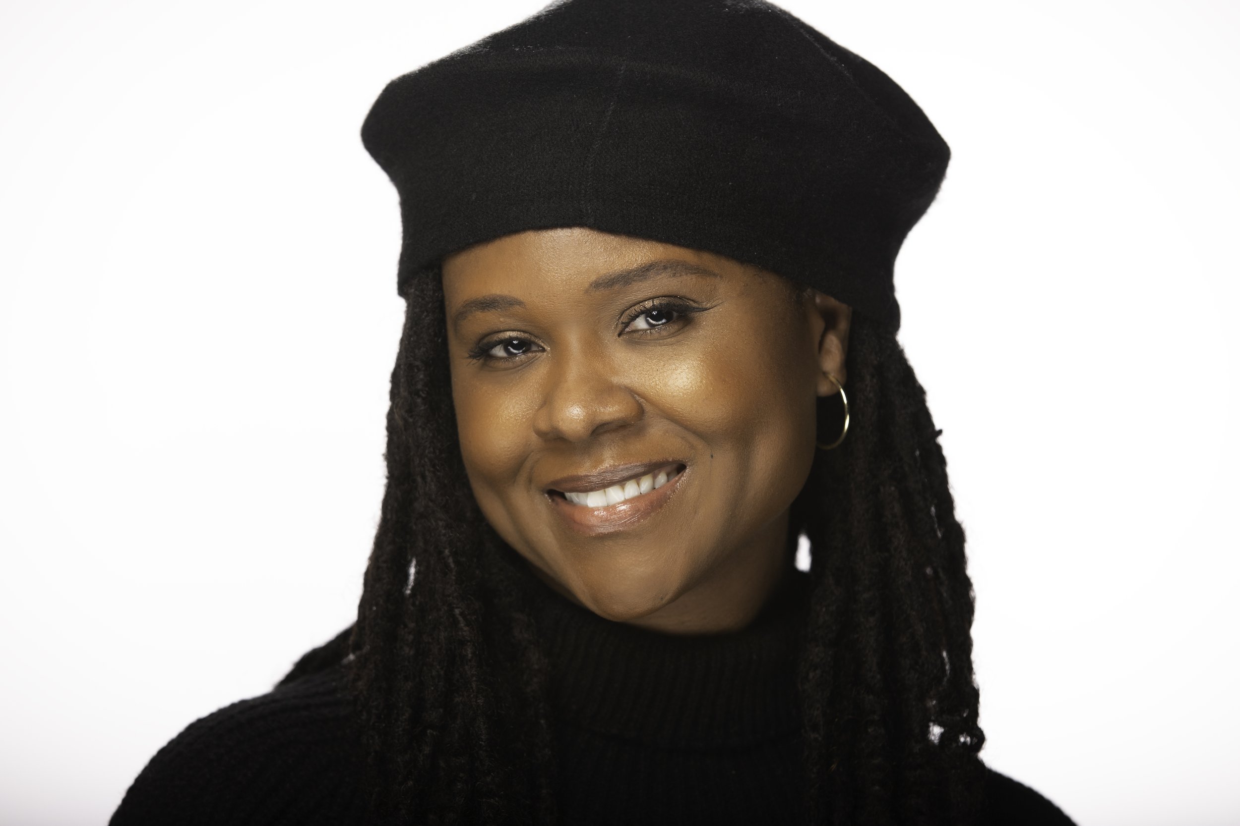 A woman smiling, wearing a black beret and black turtleneck sweater, with gold hoop earrings and long dreadlocks.