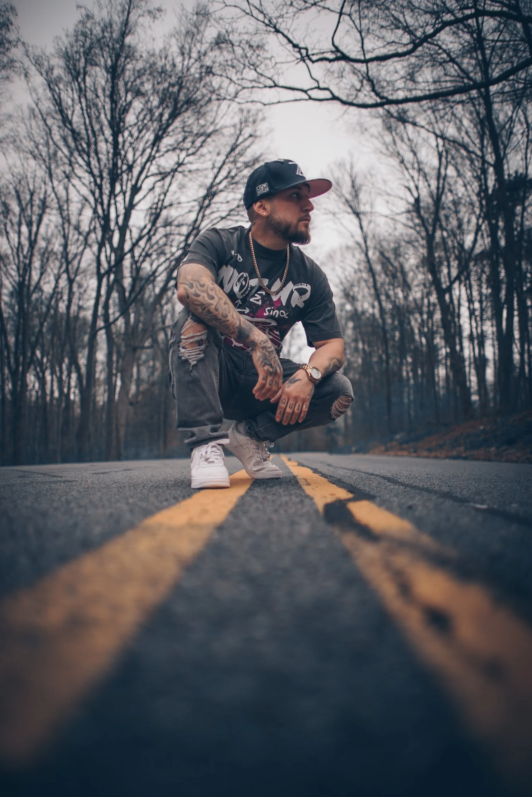 A man crouching in the middle of a two-lane road surrounded by trees with bare branches, wearing casual streetwear including a cap, graphic t-shirt, ripped jeans, and sneakers.