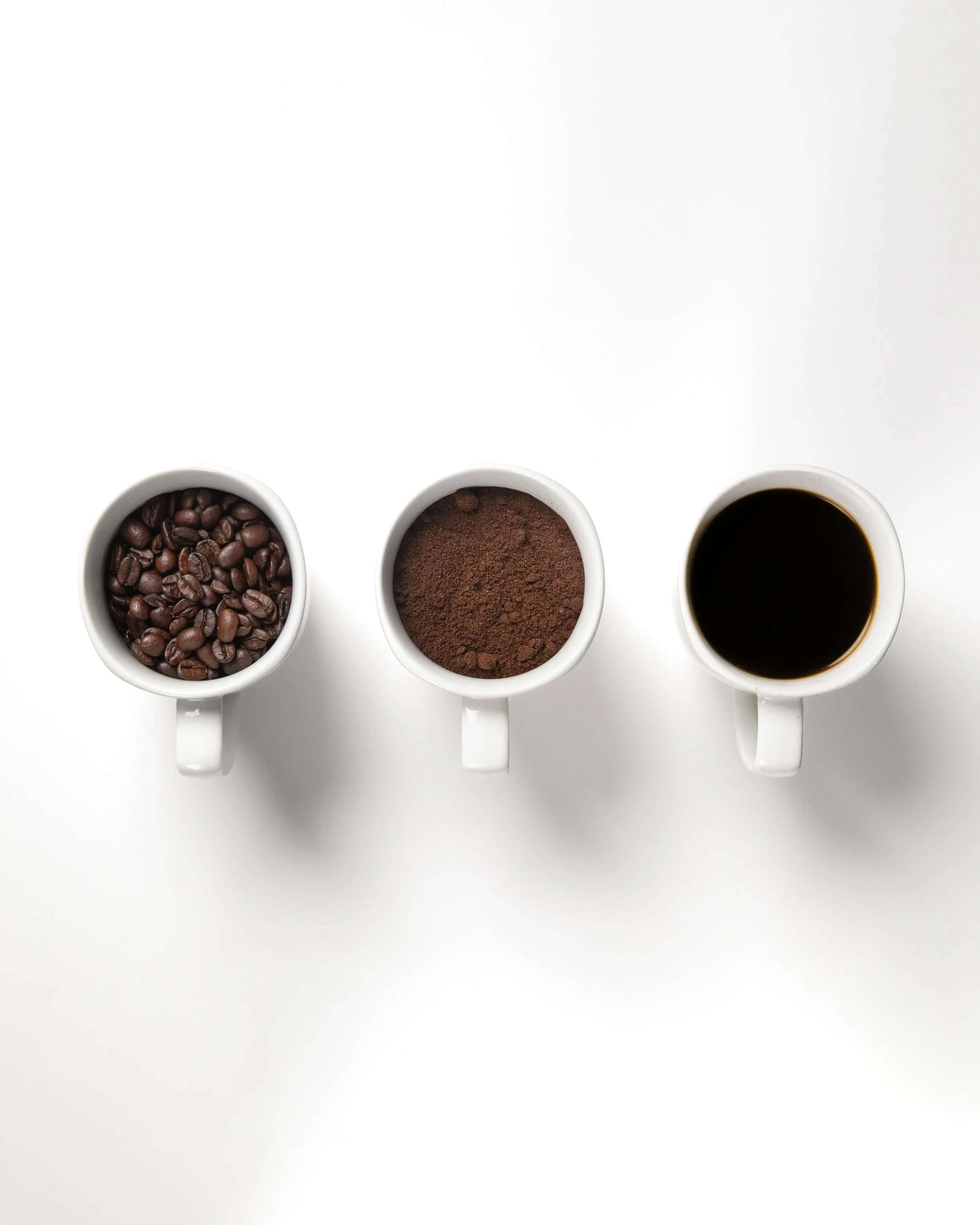 Three white coffee mugs on a white surface, containing coffee beans, ground coffee, and black coffee respectively.
