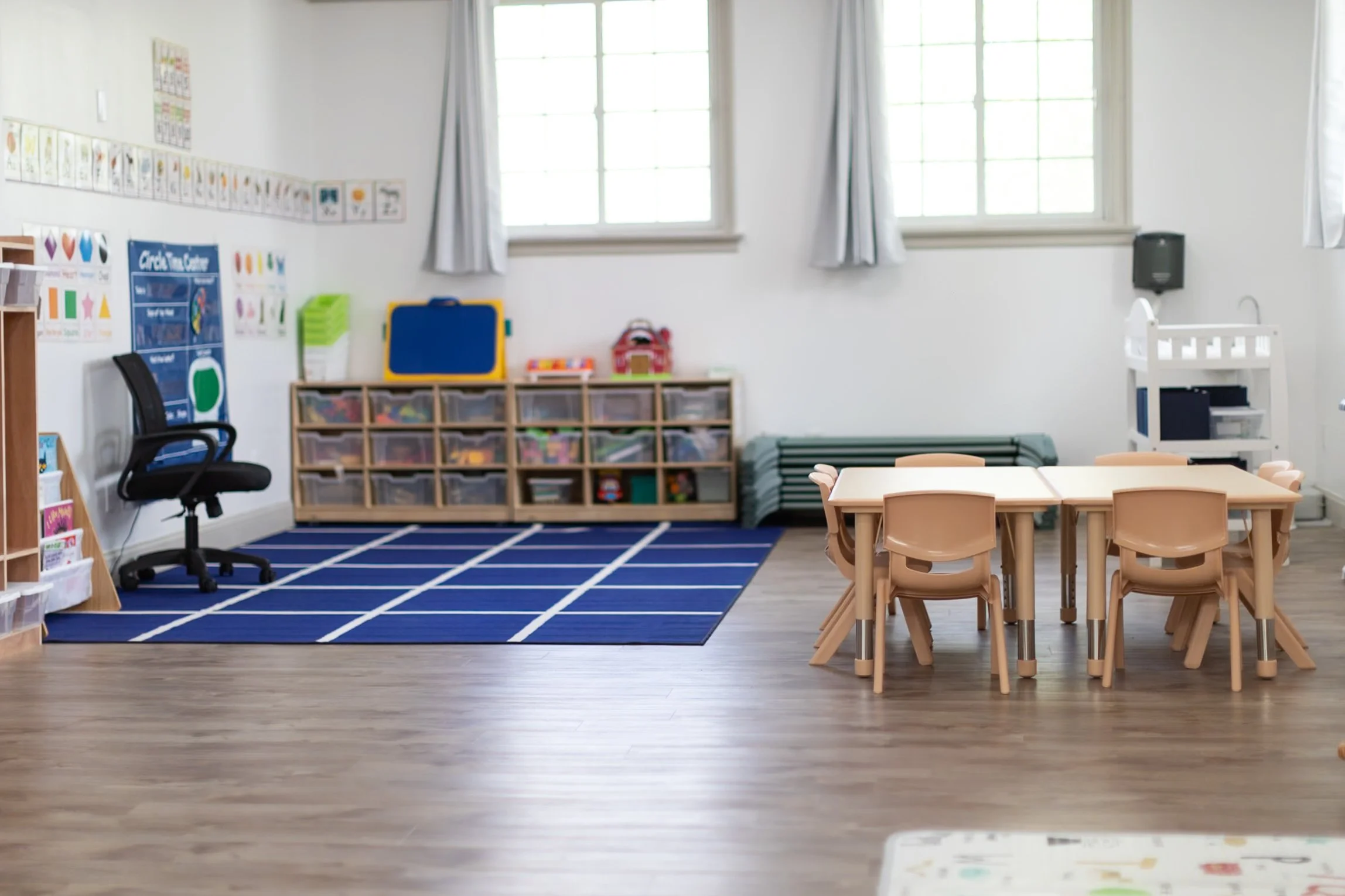 A classroom with light wood floors, white walls, and large windows. There is a blue rug with white grid lines, a black office chair, storage cubbies filled with toys and supplies, and a small table with beige chairs.