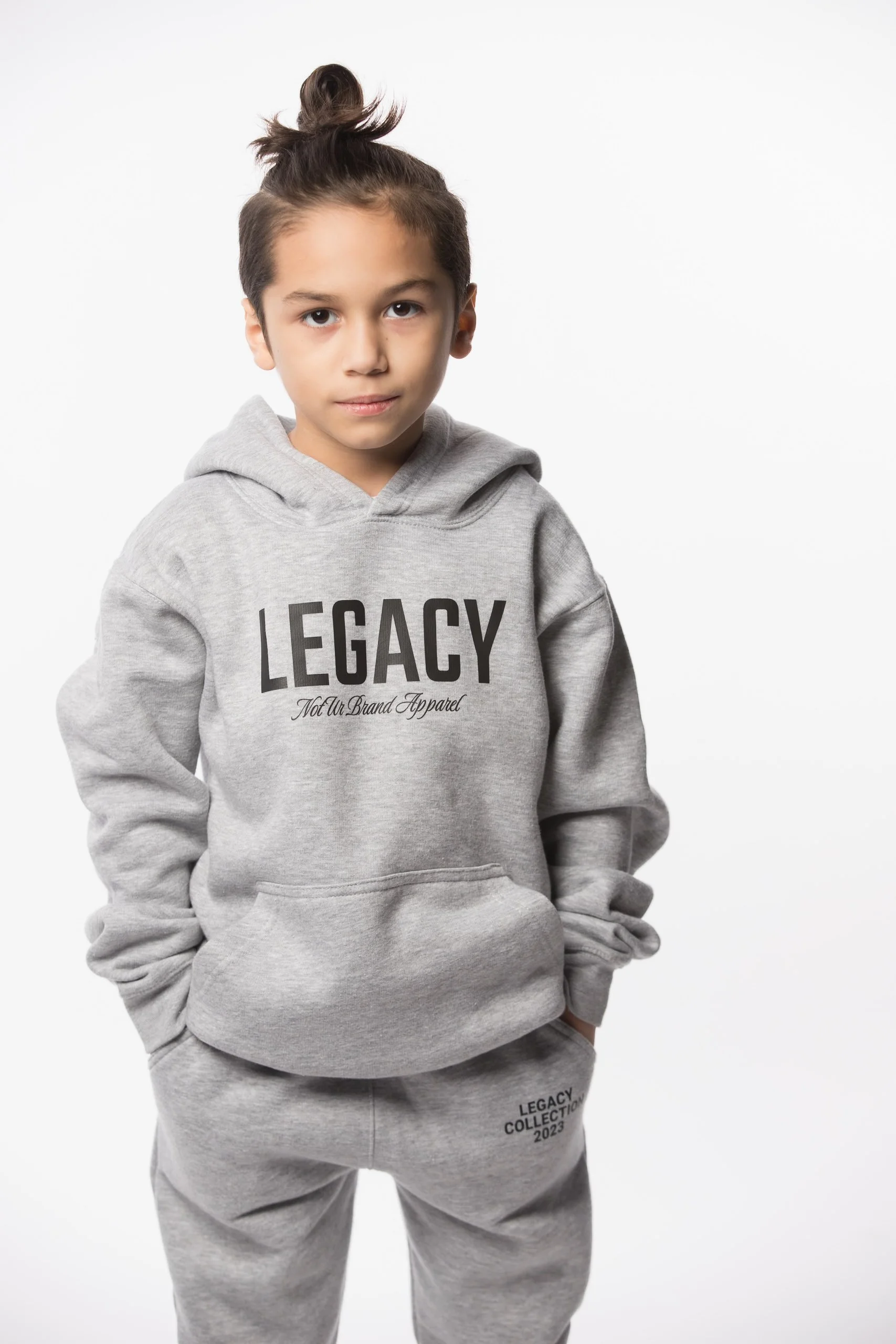 A young boy with dark hair in a top knot wearing a light gray hoodie and matching sweatpants with the word "LEGACY" printed on the front and "LEGACY COLLECTION 2023" on the pants, standing against a plain white background.
