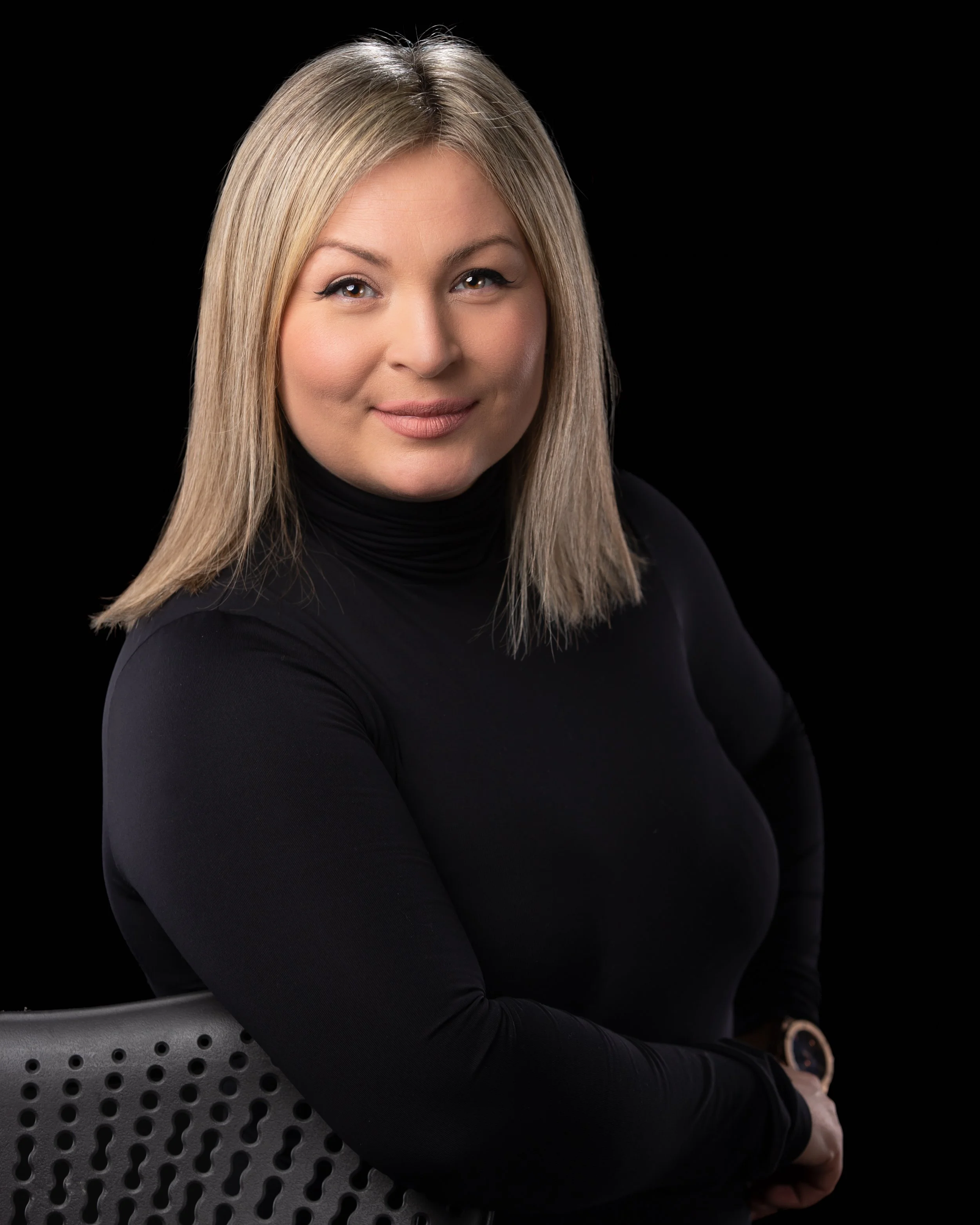 Portrait of a blonde woman wearing a black turtleneck, smiling, seated on a modern perforated chair with a black background.