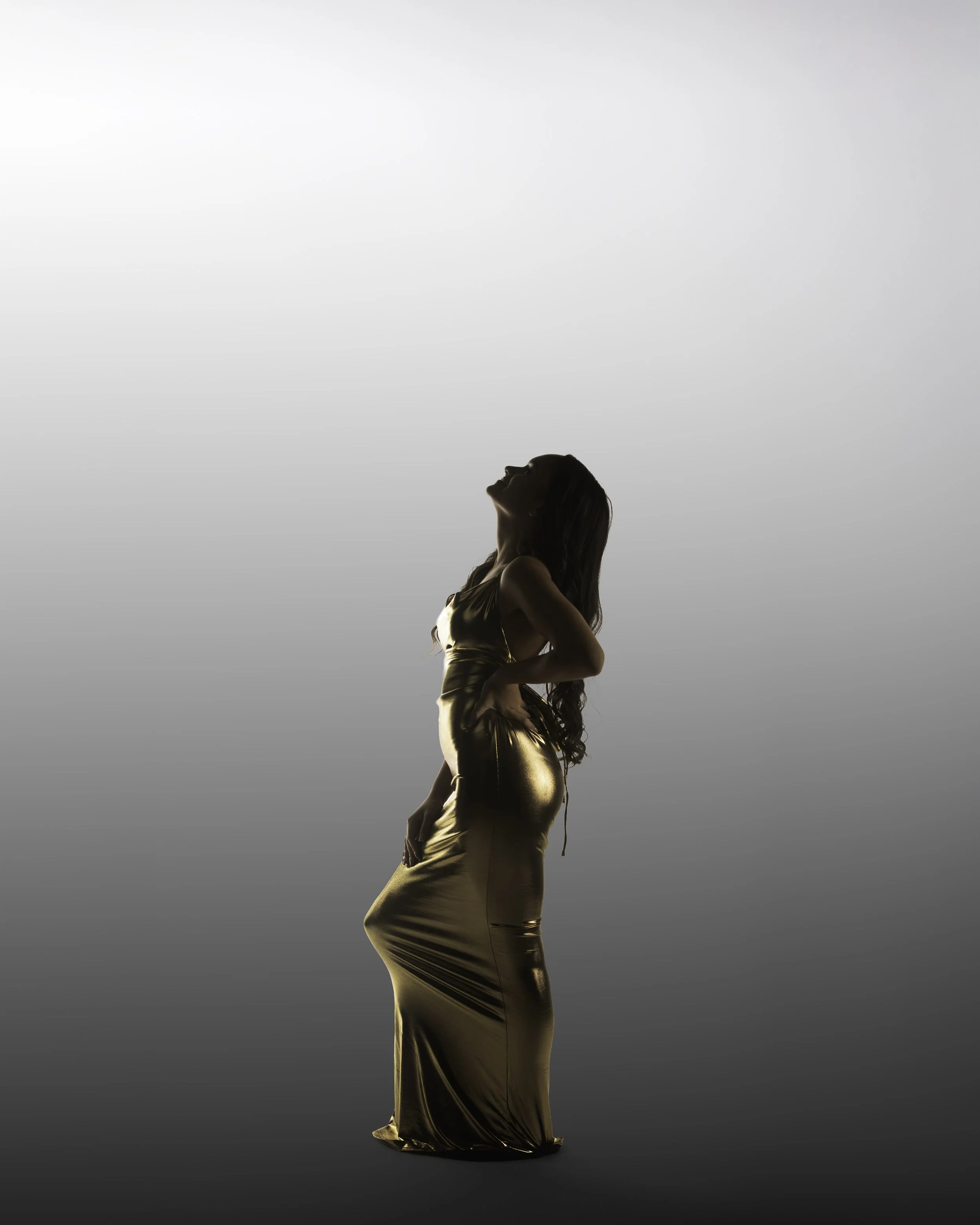 A woman in a shiny gold dress stands with her face tilted upward and hands on her hips against a gradient background.