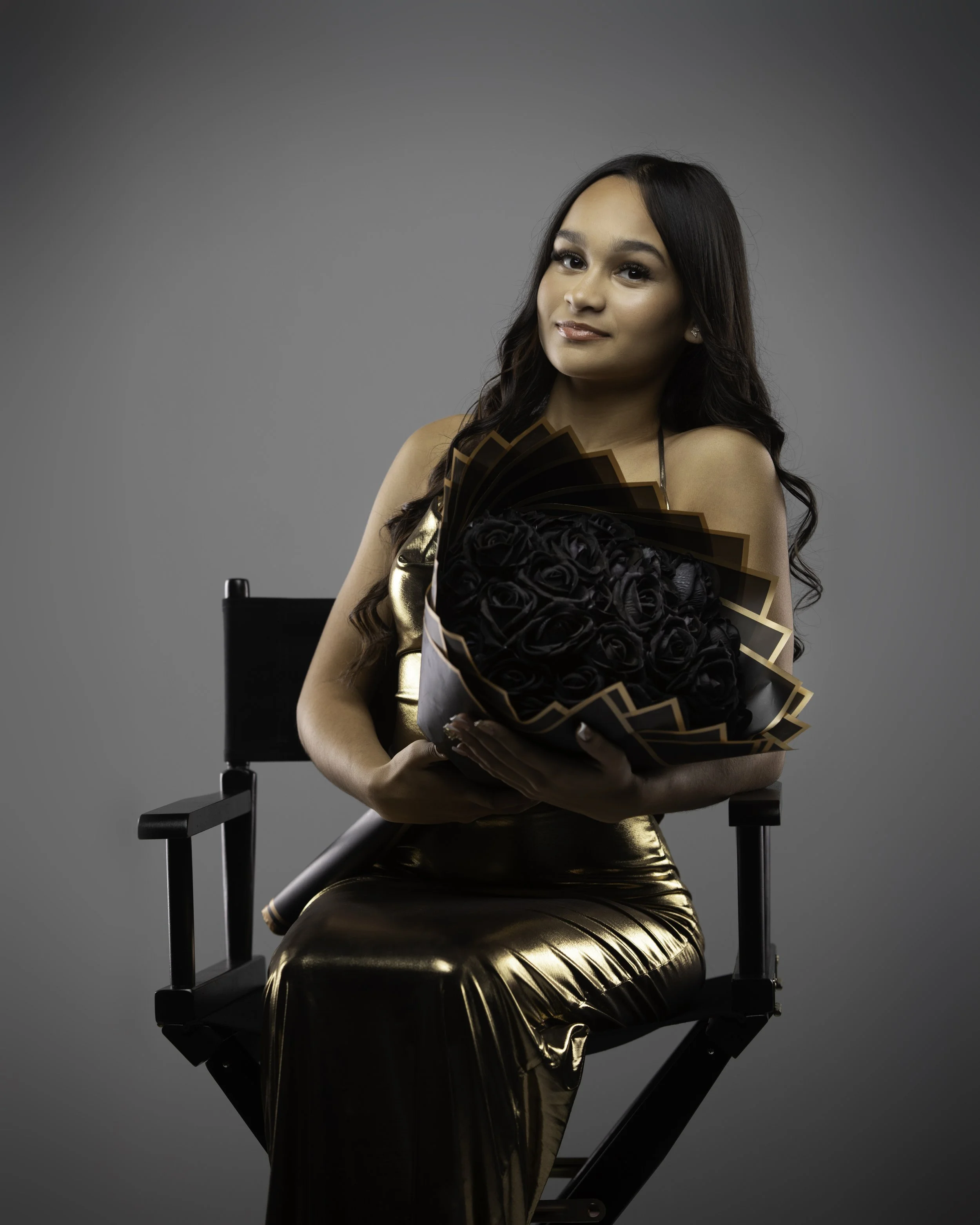 Woman in gold dress sitting on director's chair, holding a bouquet of black roses, against gray background.