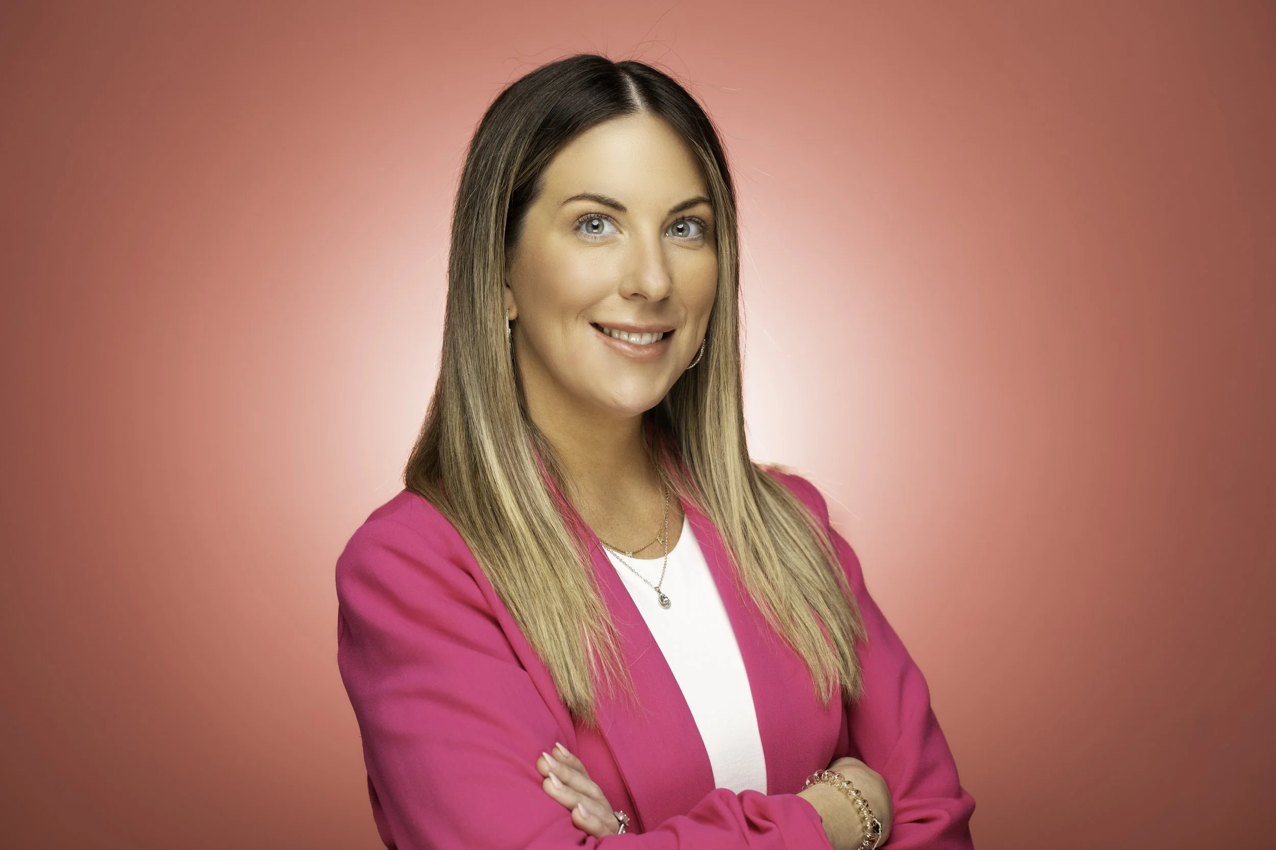 A woman with long blonde hair smiling, wearing a bright pink blazer over a white top, standing against a pink background.