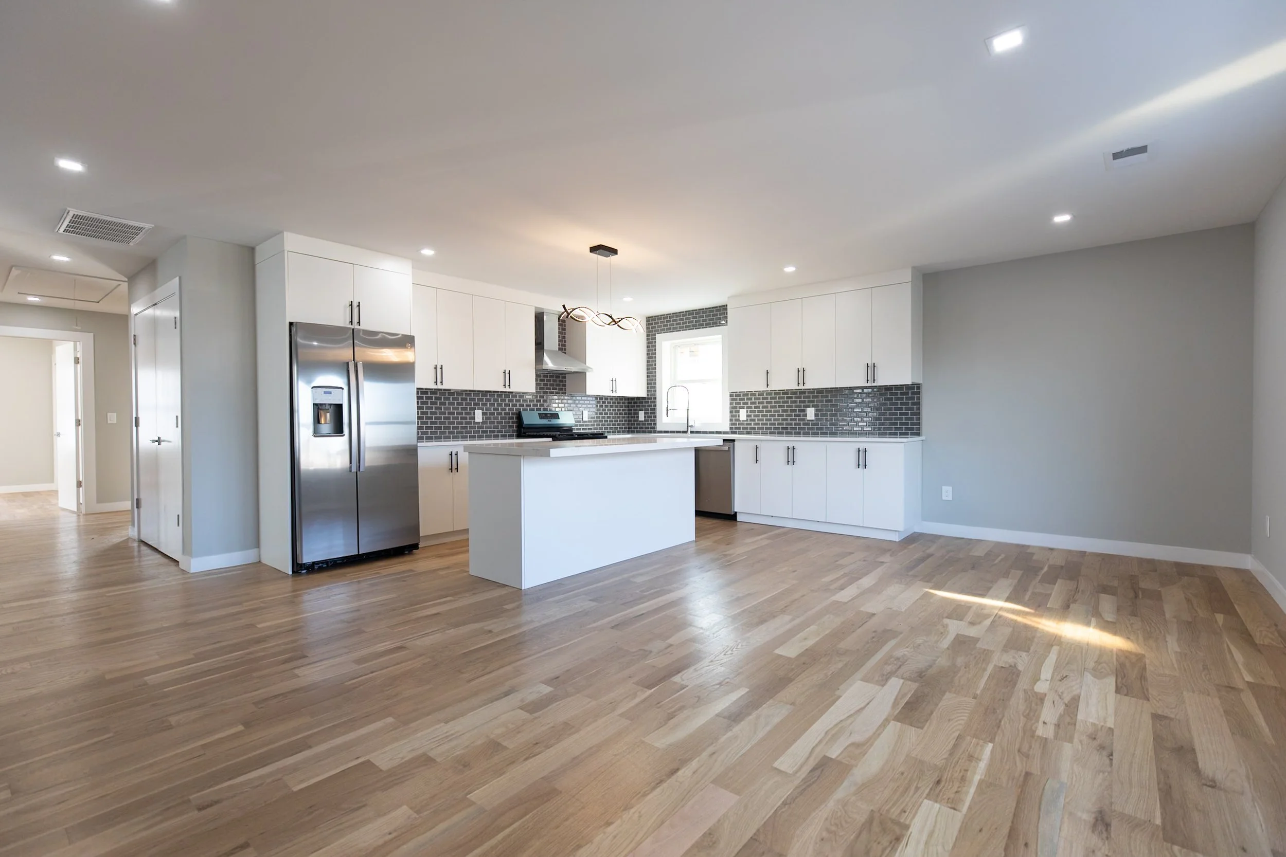 Modern kitchen with white cabinets, stainless steel refrigerator, gray backsplash, island, hardwood floors, and natural light from window