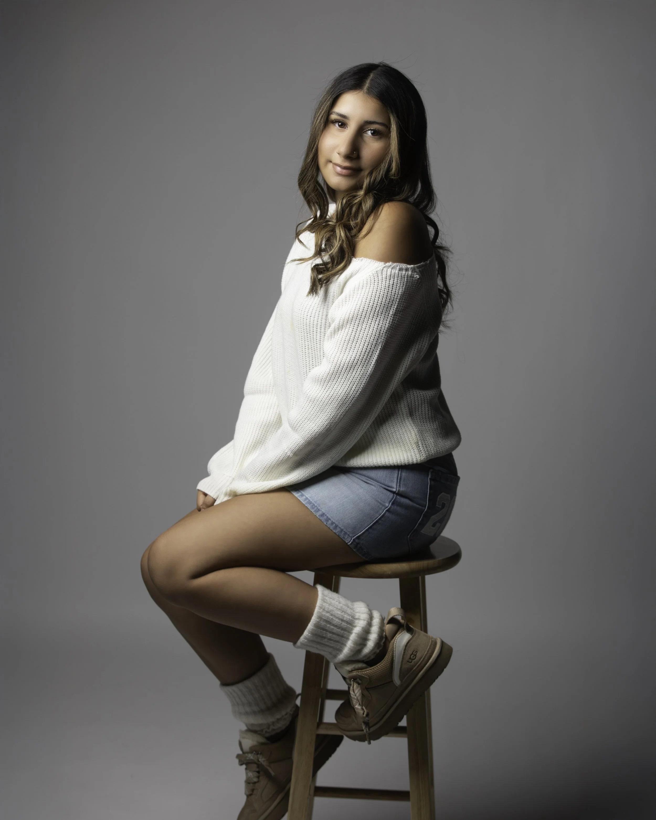 A young woman with long brown hair, sitting on a wooden stool in a casual pose, wearing a white off-shoulder sweater, denim shorts, tan boots, and beige socks, against a plain gray background.