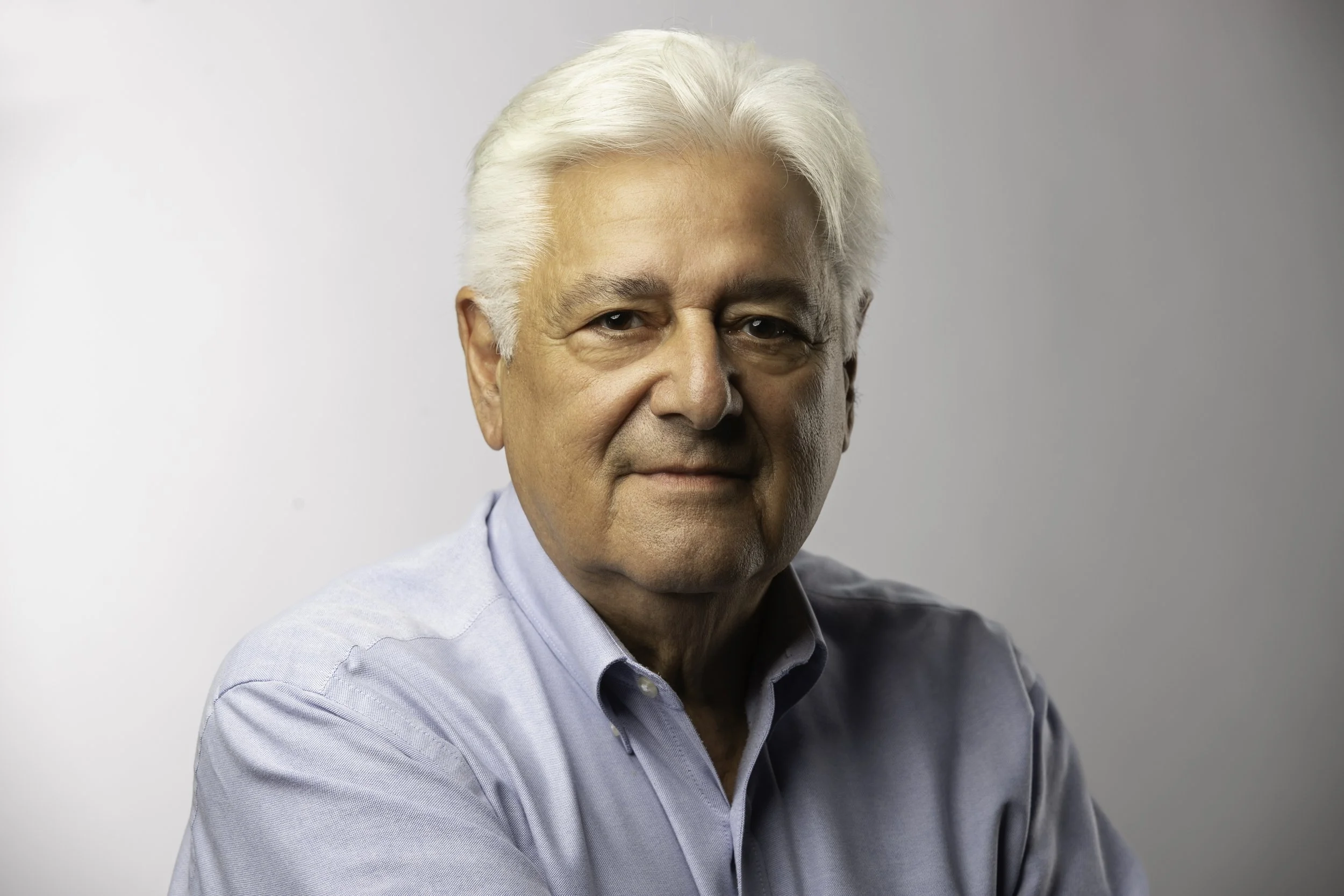 Portrait of an elderly man with white hair wearing a light blue shirt against a plain background.