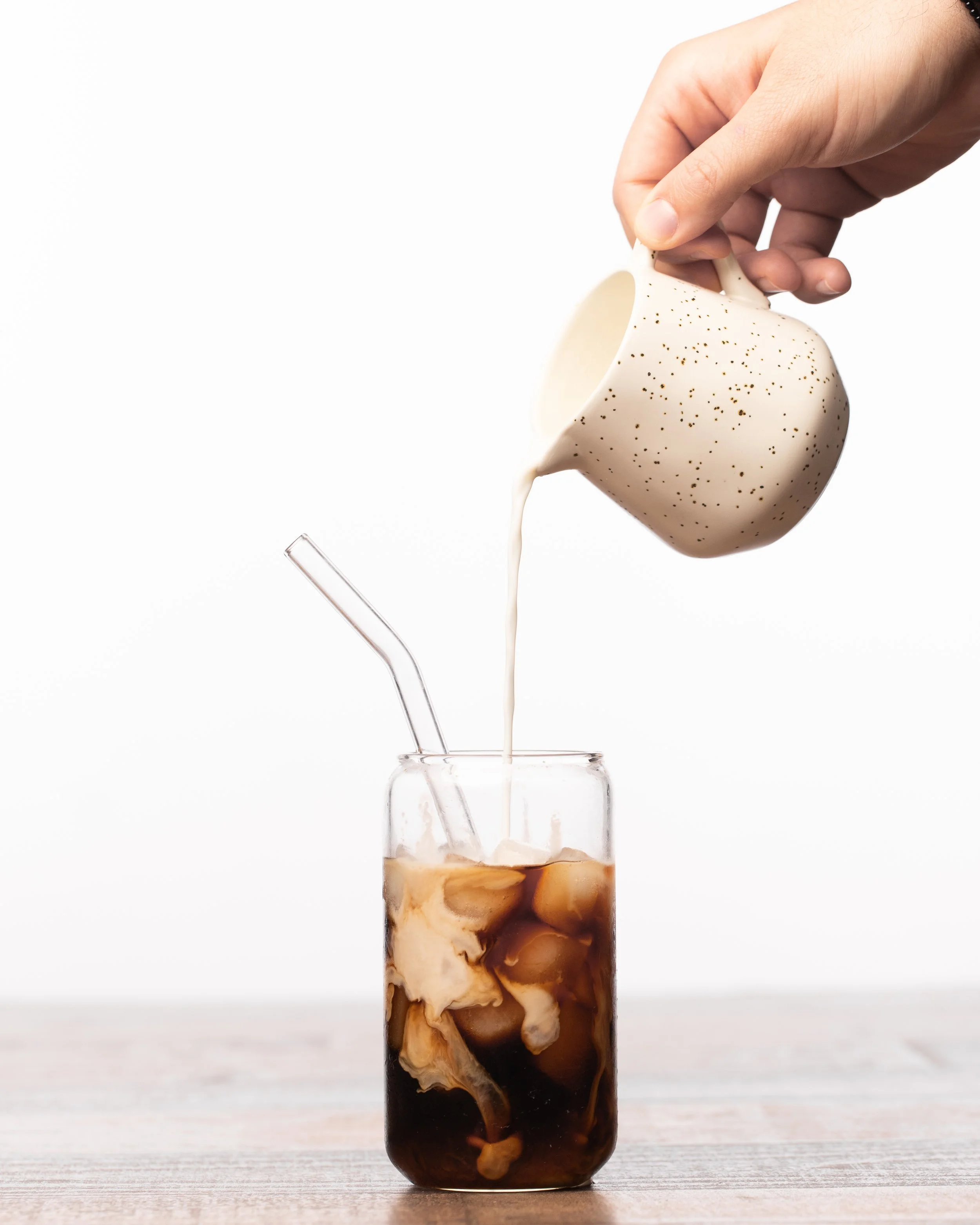 Hand pouring cream into a glass of iced coffee with a straw on a wooden surface