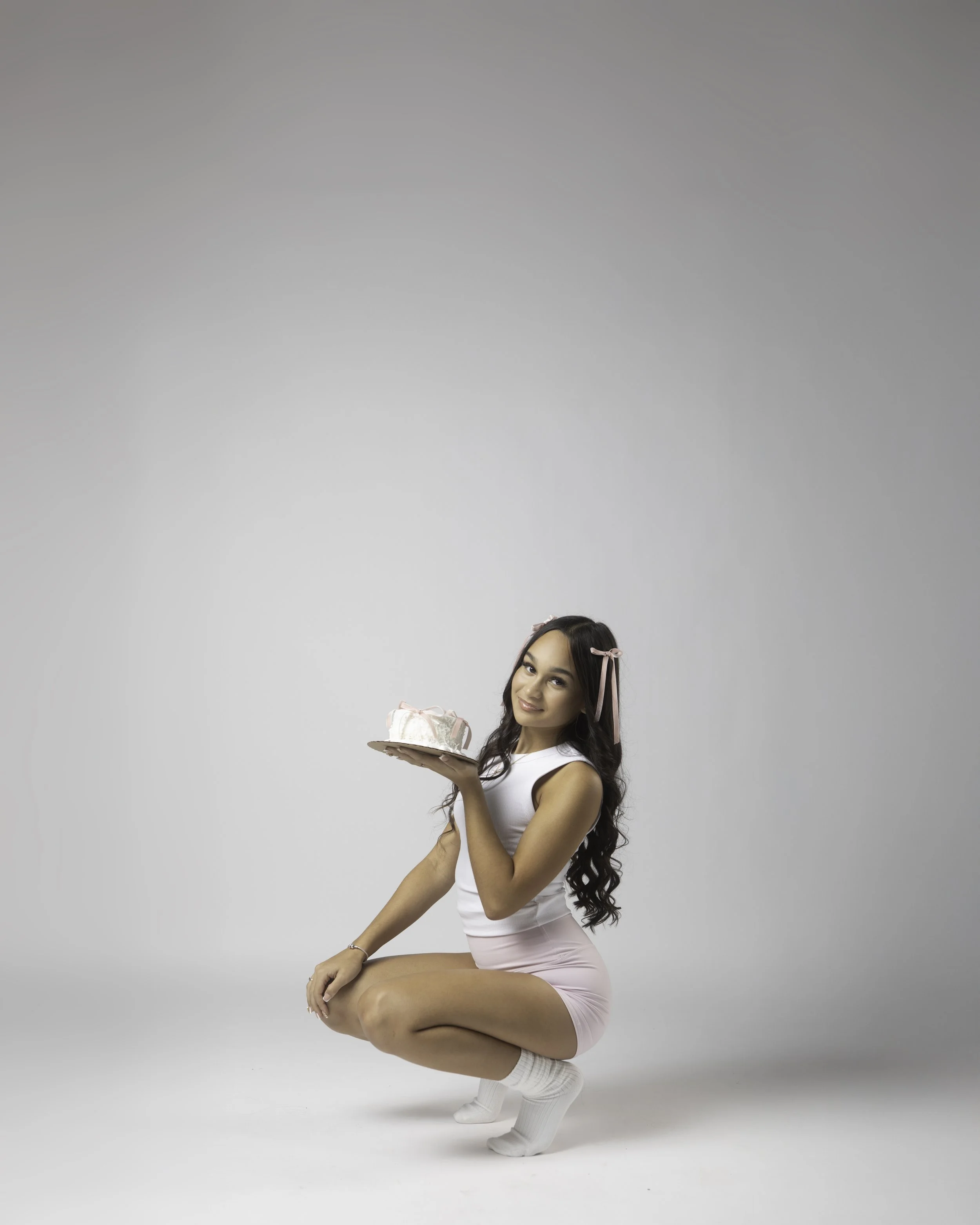 A young woman with long dark hair kneeling on a white floor, wearing a white top, pink shorts, and white socks, holding a cake with pink and white frosting, smiling.