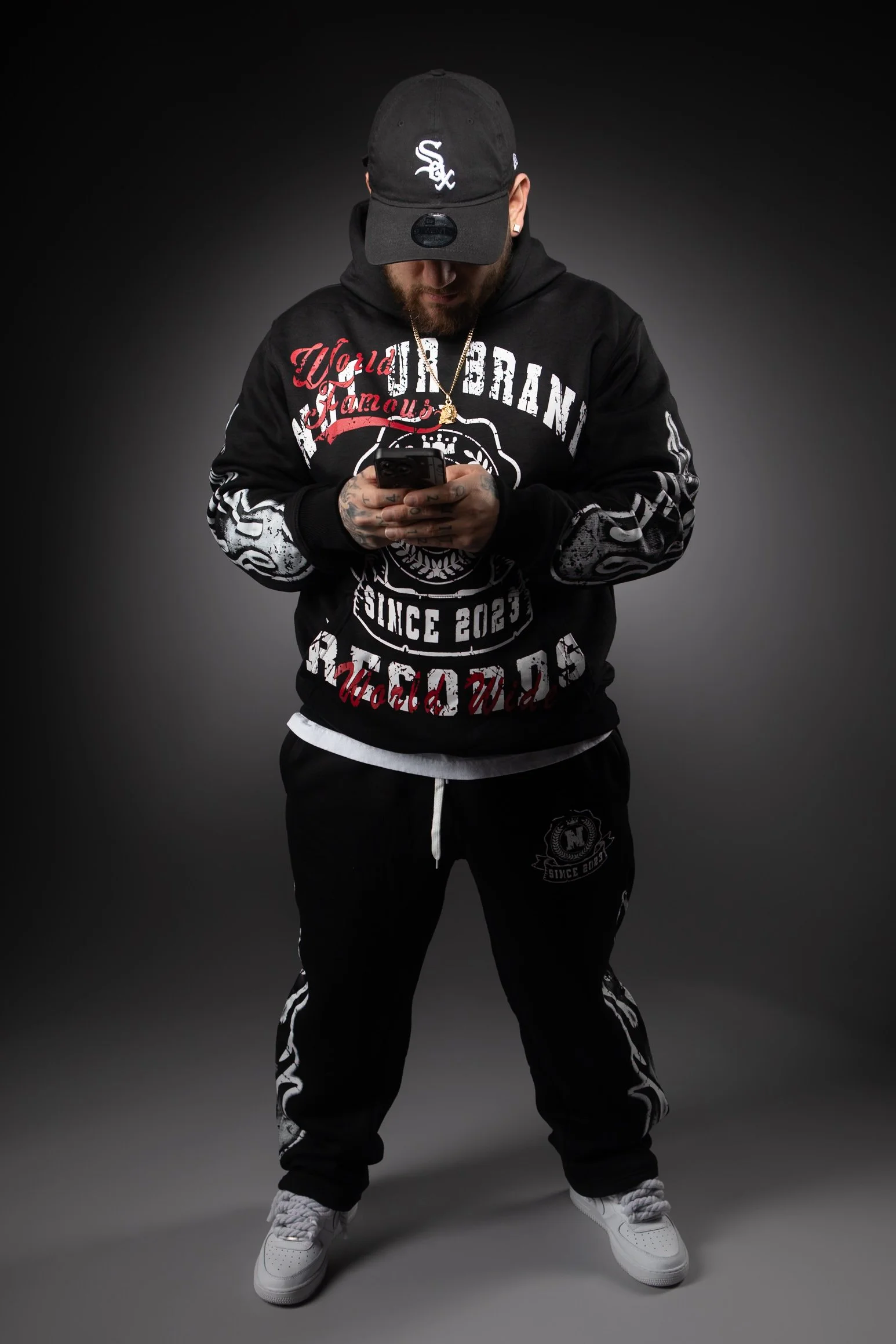 A man wearing a black hoodie, black sweatpants, white sneakers, and a black baseball cap with the Chicago White Sox logo, is looking down at his phone against a dark background.