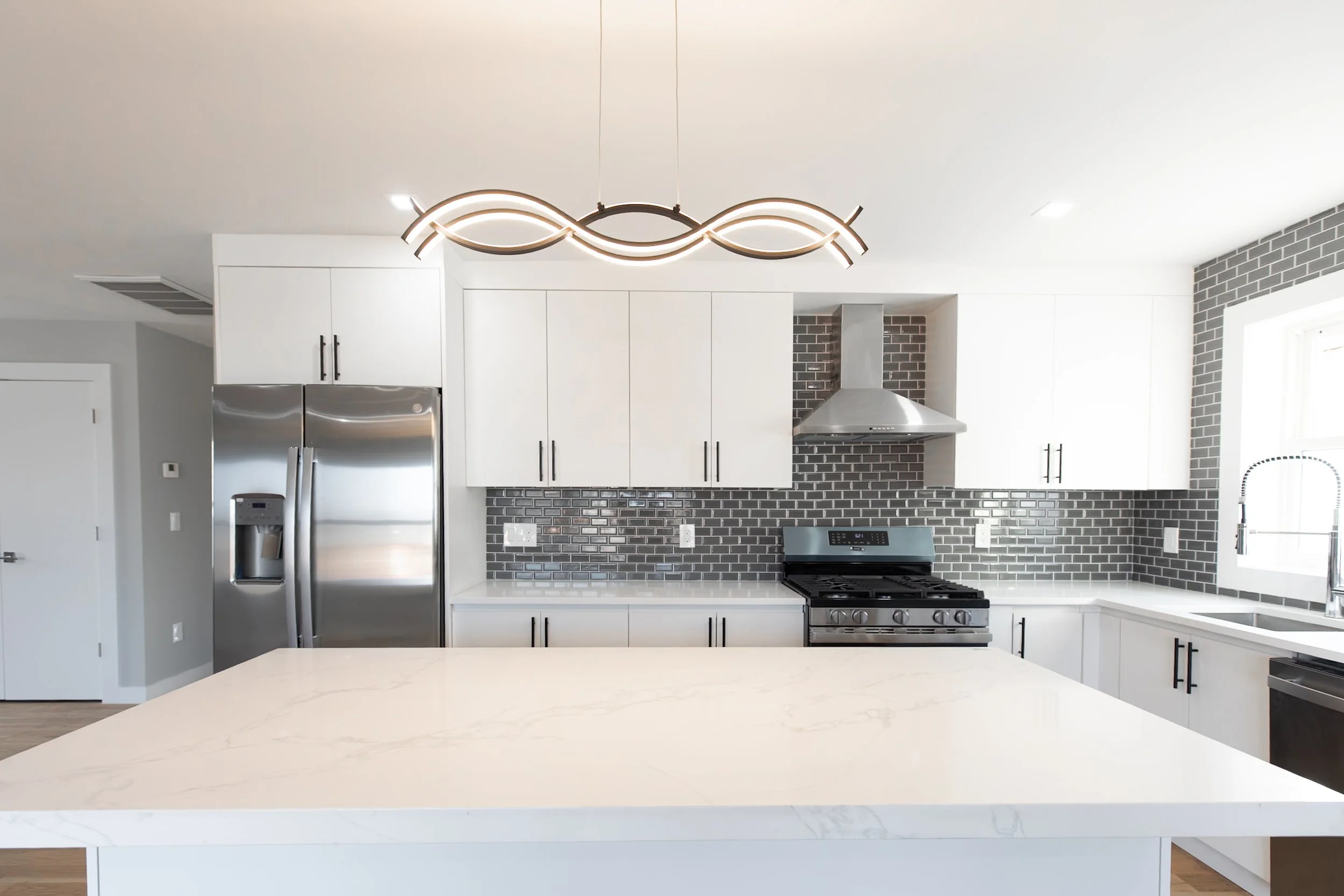 Modern kitchen with white cabinets, stainless steel refrigerator and stove, gray brick backsplash, large white island, and contemporary light fixture.