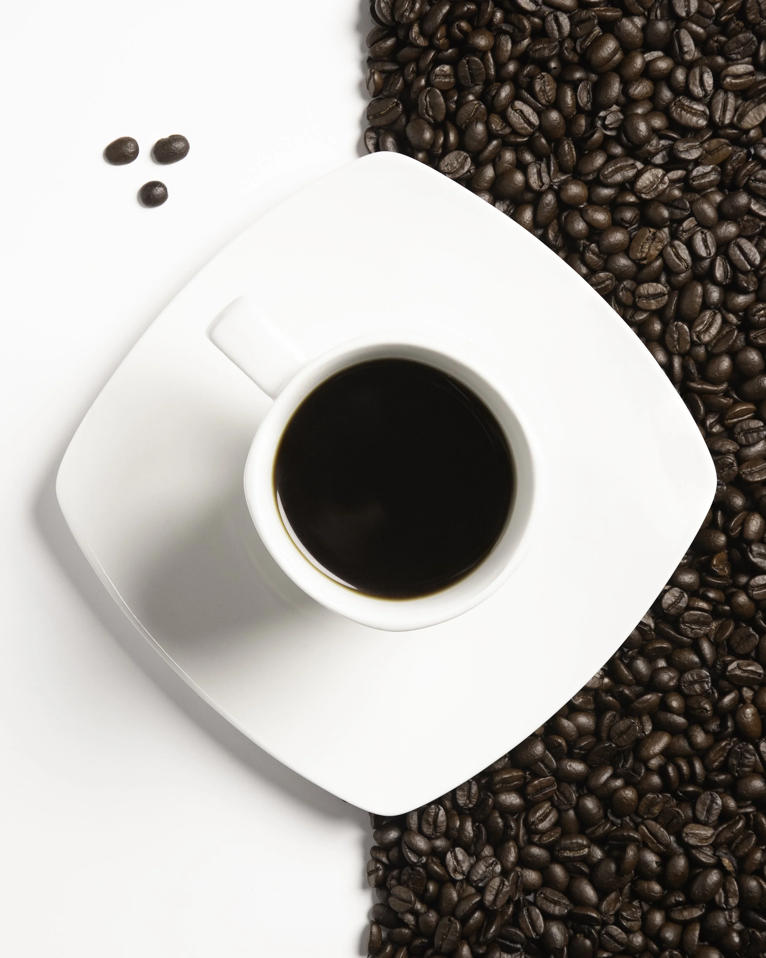 A top-down view of a white cup filled with black coffee on a white saucer, surrounded by scattered coffee beans on a white surface.