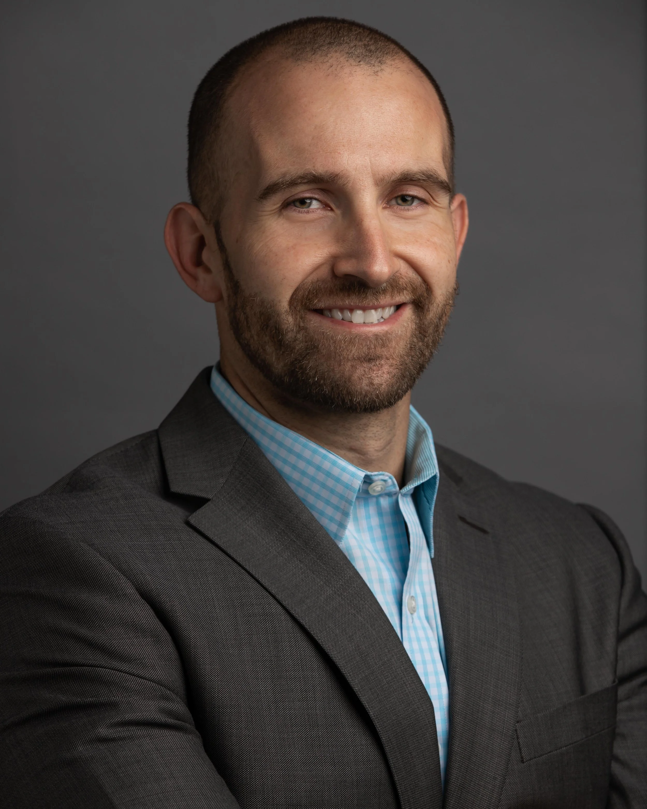 A professional headshot of a smiling man with a beard, wearing a dark suit and blue checkered shirt, against a gray background.