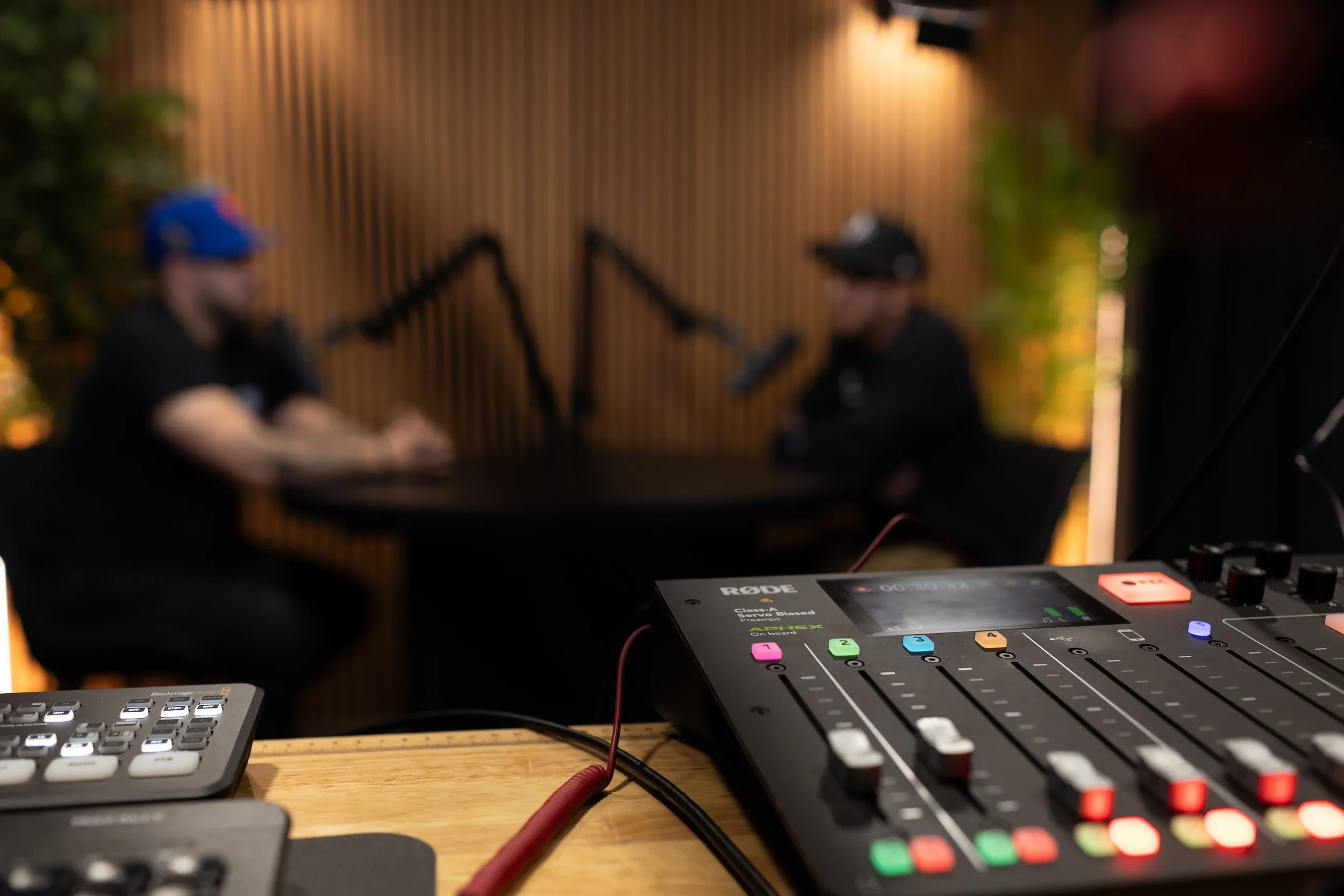 Audio mixing board in foreground with blurred two people sitting and speaking into microphones in background.