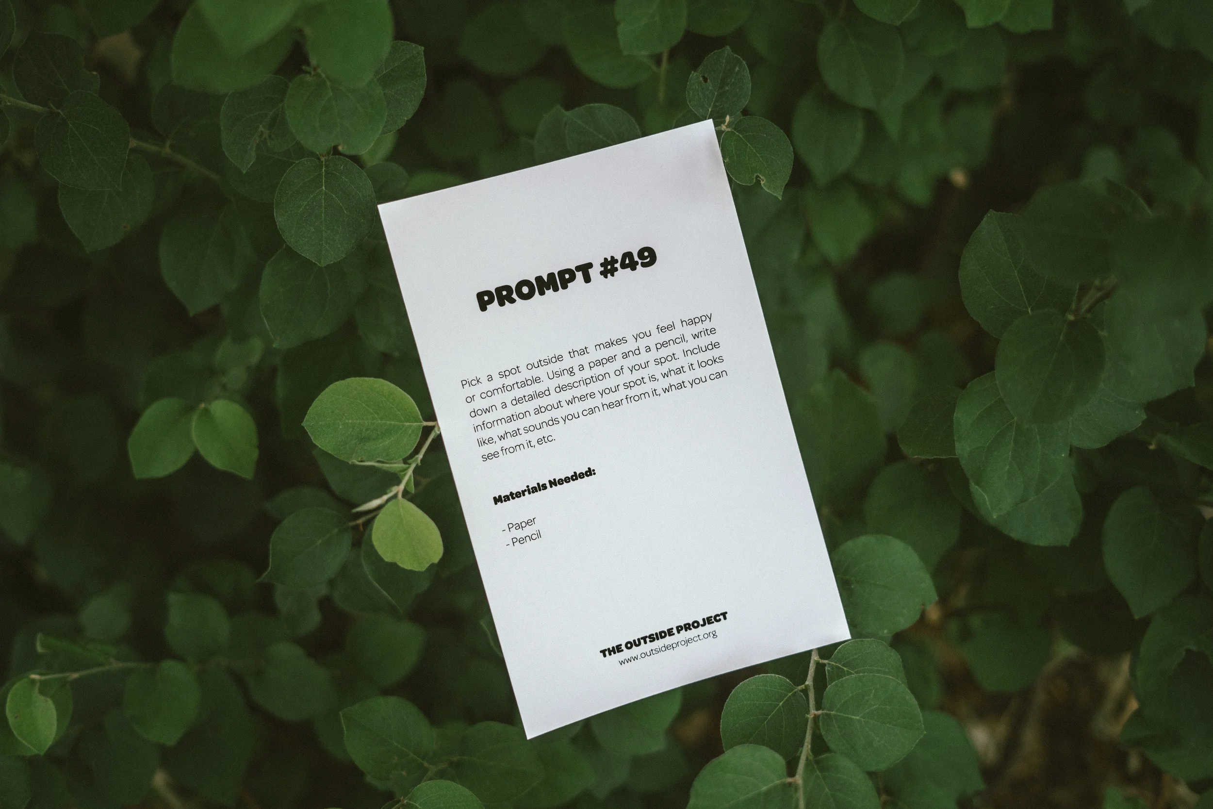 A white paper titled 'PROMPT #49' attached to green leafy plants, with instructions to pick a happy or comfortable spot outside, describe the scene, and note sounds, with materials needed listed as paper and pencil, and a footer with a website URL.