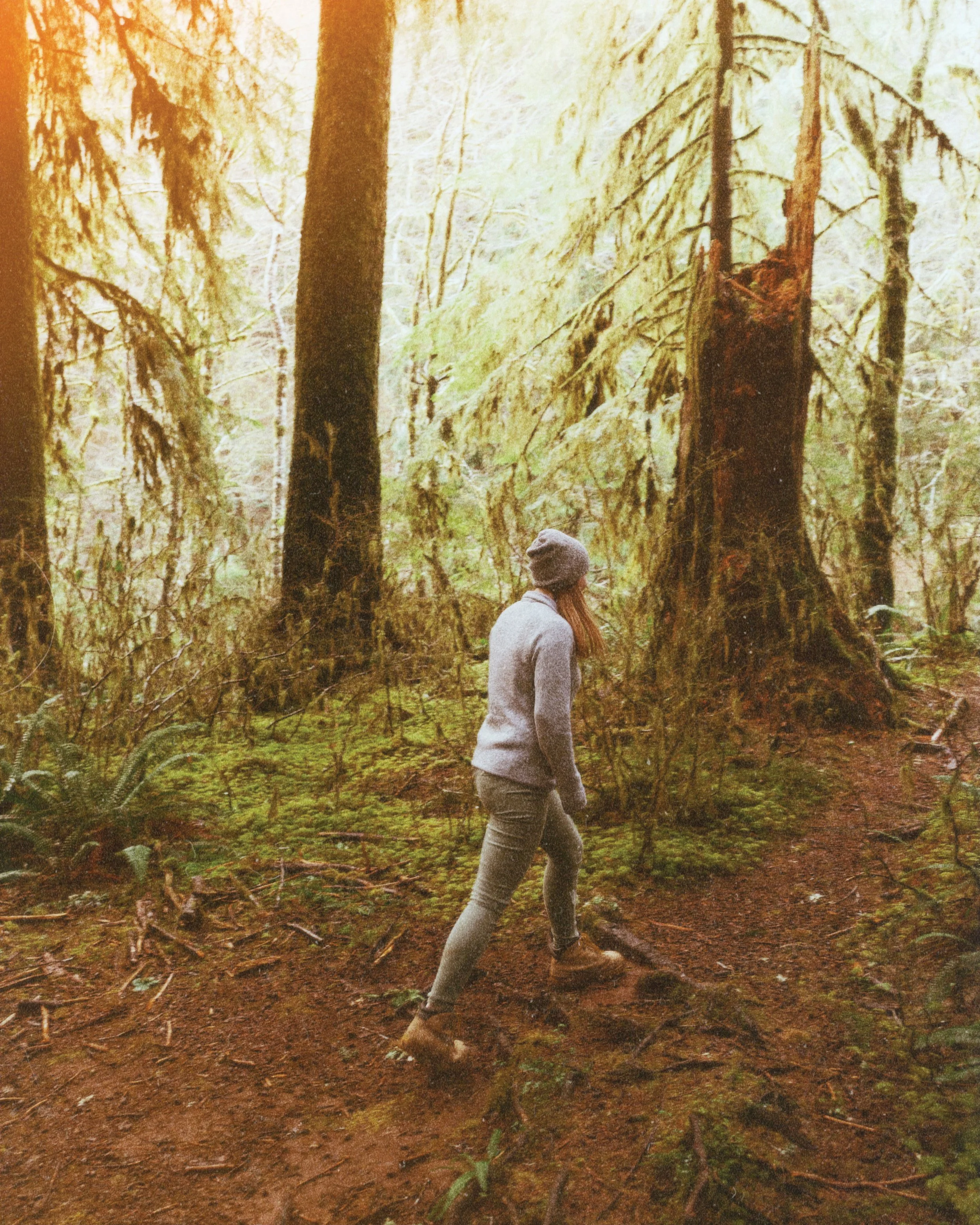 A woman walking on a forest trail surrounded by tall trees and lush green foliage, wearing a gray sweater, gray pants, and a beanie.