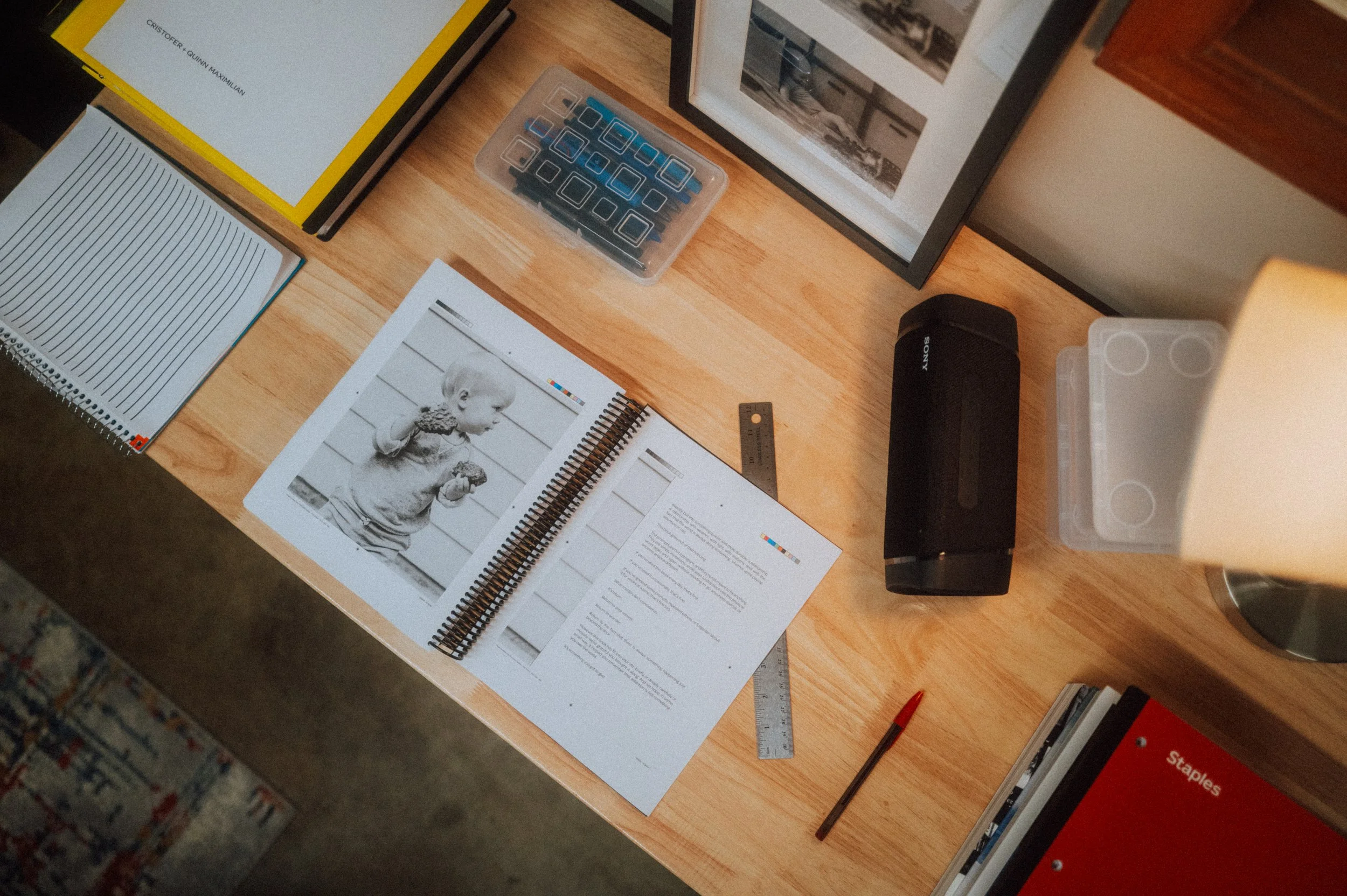A top-down view of a wooden desk with various office items including notebooks, a ruler, a pen, a photo album, framed pictures, a black portable speaker, and storage boxes.
