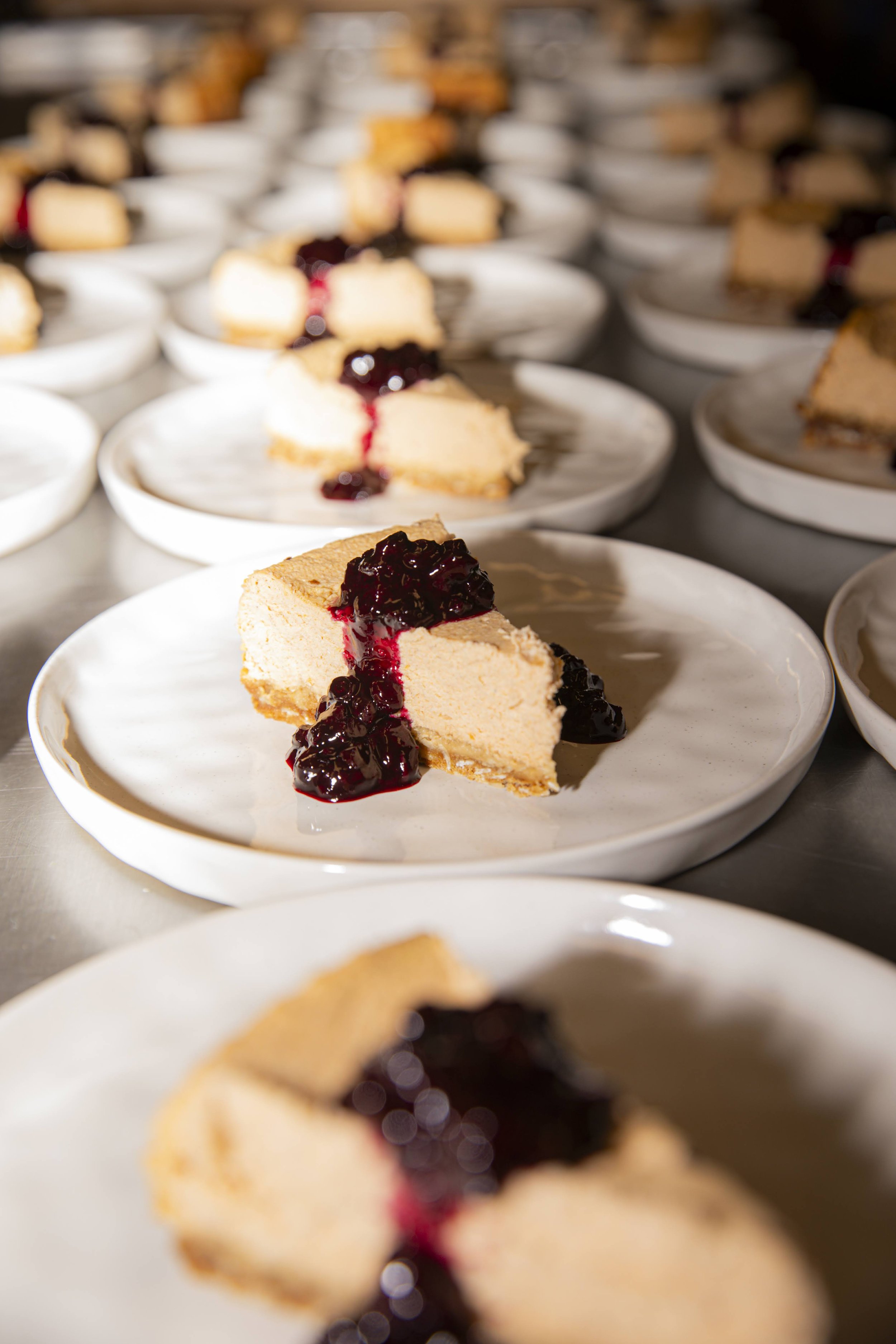 Multiple plates with slices of cheesecake topped with berry syrup, arranged in rows on a kitchen counter.