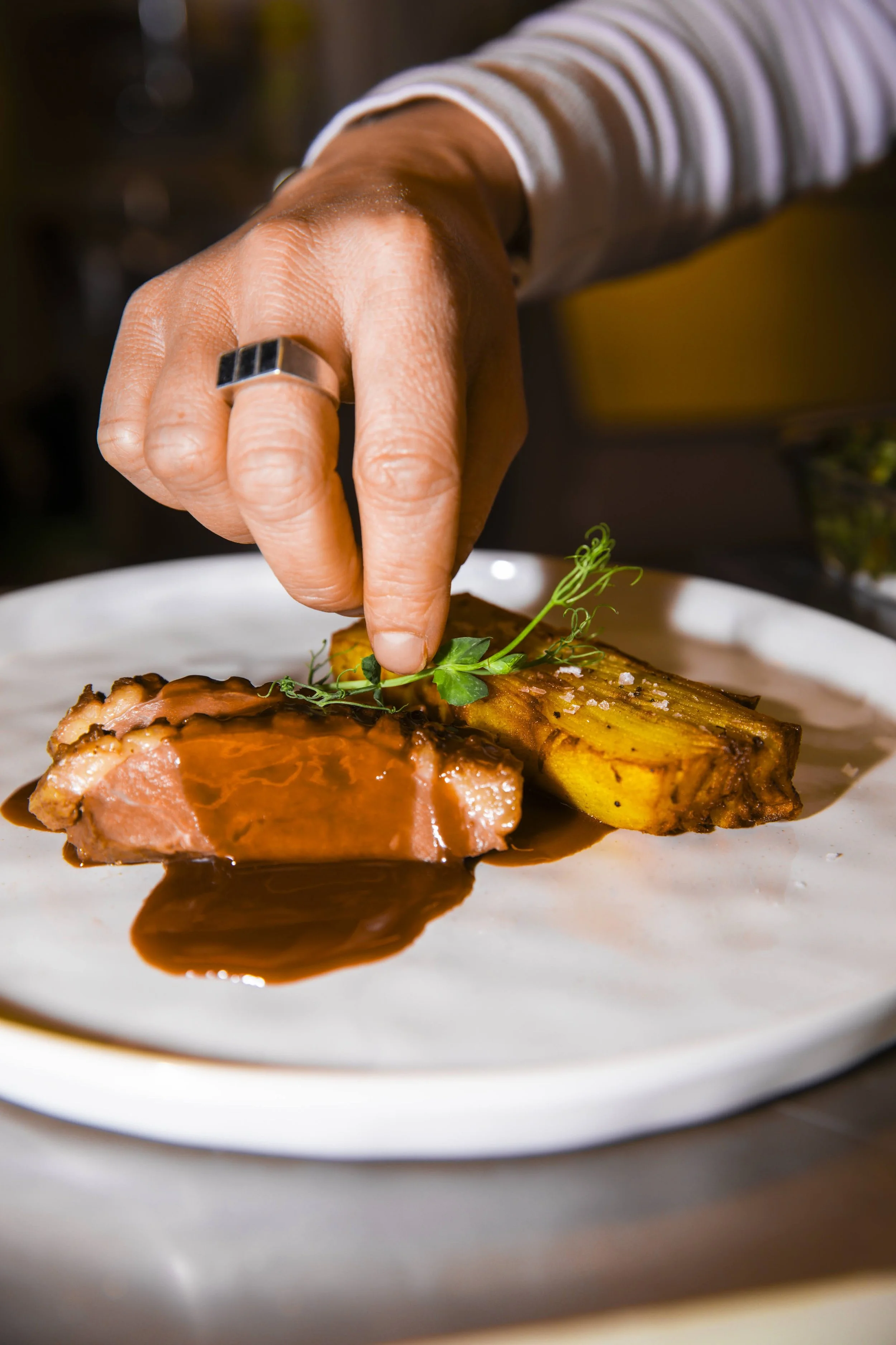 Close-up of a hand with a ring, gently placing a sprig of green herbs on a slice of cooked meat with sauce, on a white plate.