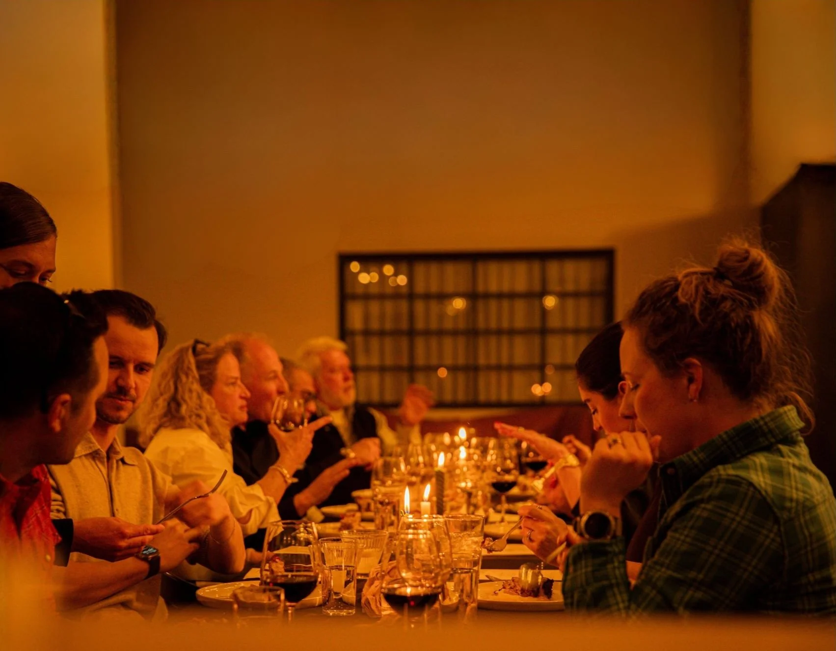 People sitting at a long dinner table in a warmly lit room, engaged in conversation and eating during a social gathering or dinner event.