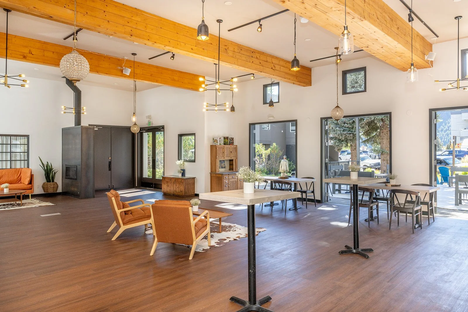 Spacious, modern room with wooden beams, large windows, and stylish furniture, including chairs, tables, and decorative plants, in a well-lit interior with hardwood floors.