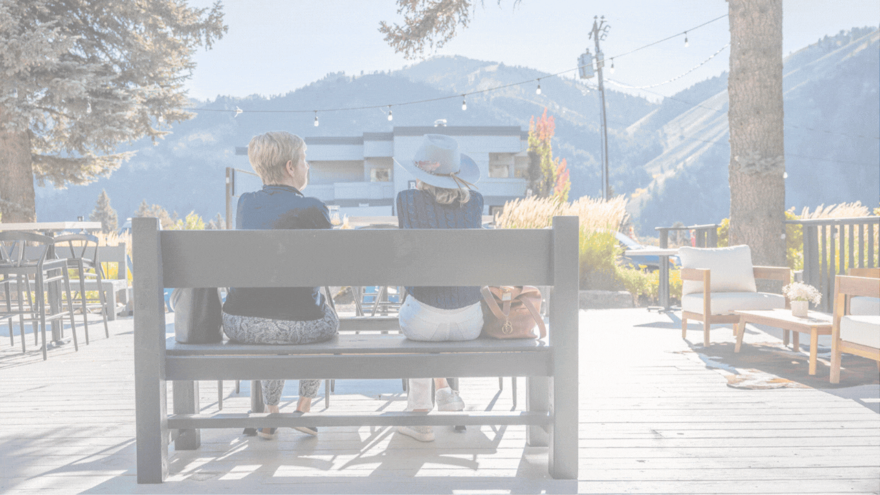 Two women with hats sit on a bench outdoors, talking with their backs to the camera, in a mountain setting with string lights overhead and trees around.