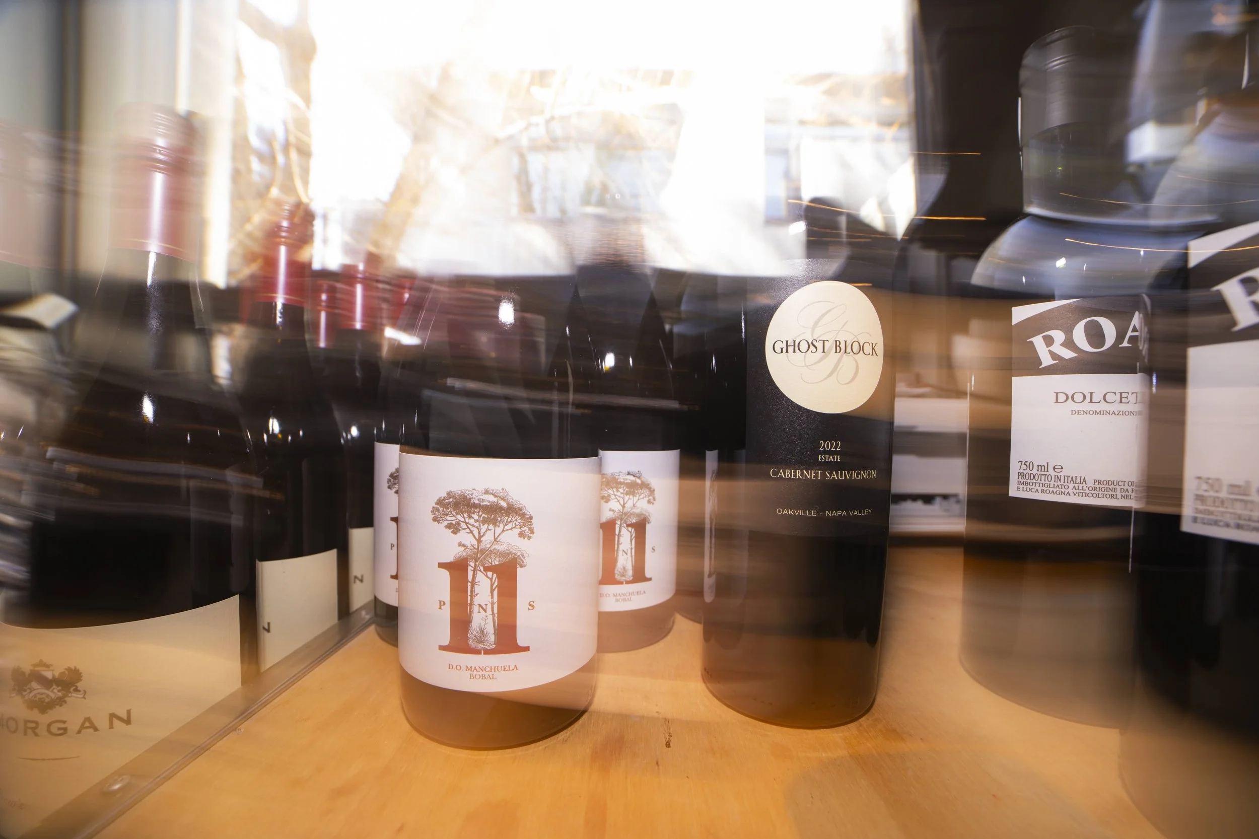 A display of wine bottles on a wooden shelf with sunlight reflecting off the glass, some bottles have labels visible including 'Ghost Block' and 'Dolan.'