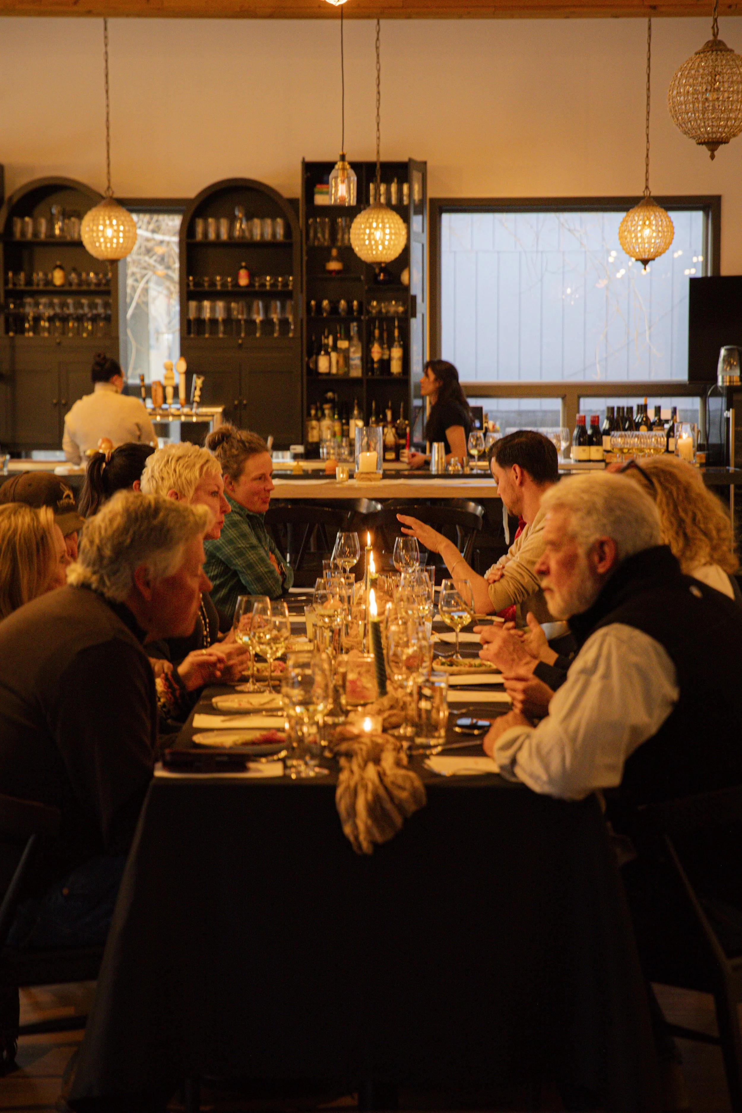 A group of people sitting at a long table in a restaurant, engaged in conversation, with wine glasses and candles on the table, and a bar in the background.