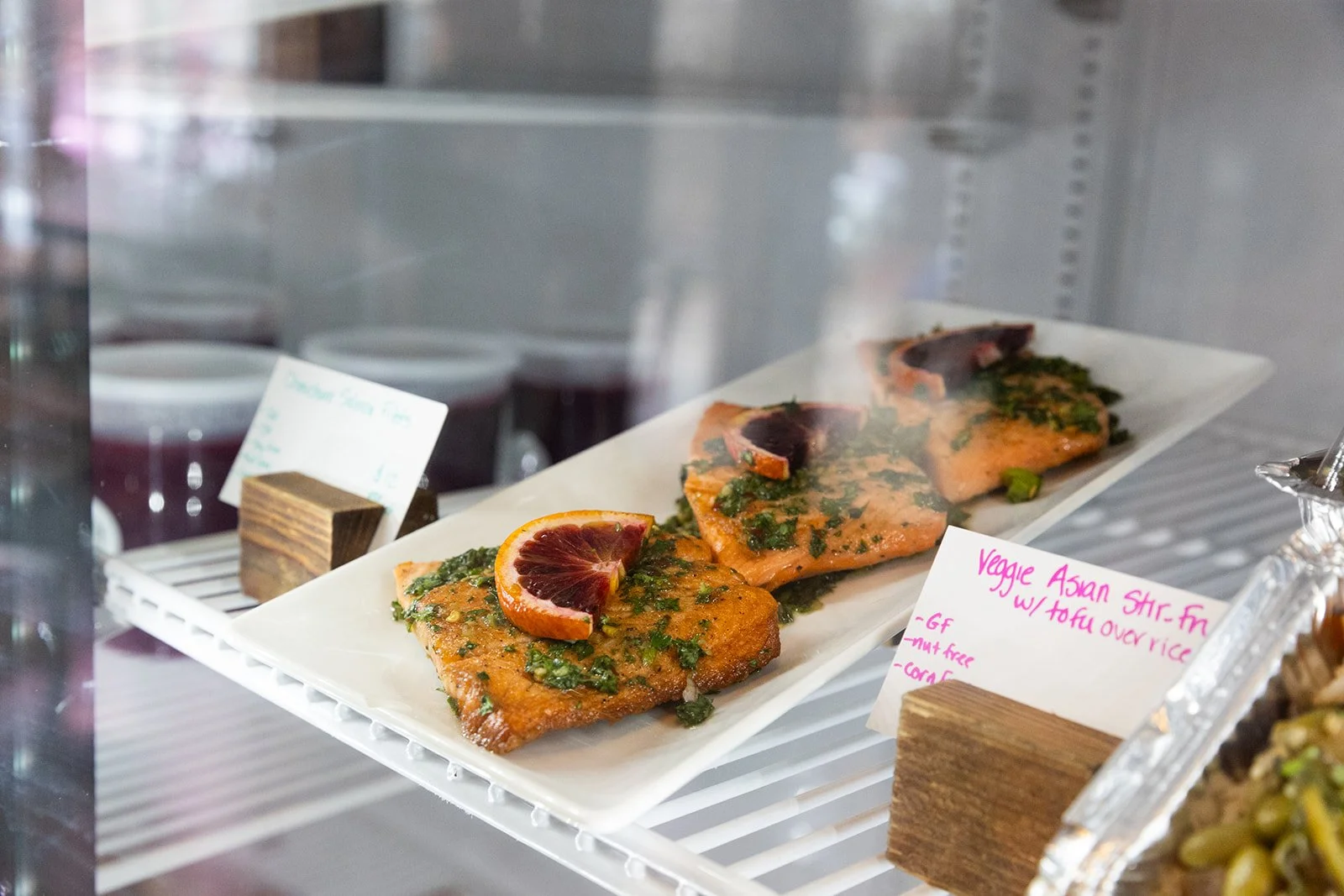 Plate of three cooked salmon fillets garnished with herbs and a slice of blood orange, inside a display case at a restaurant.