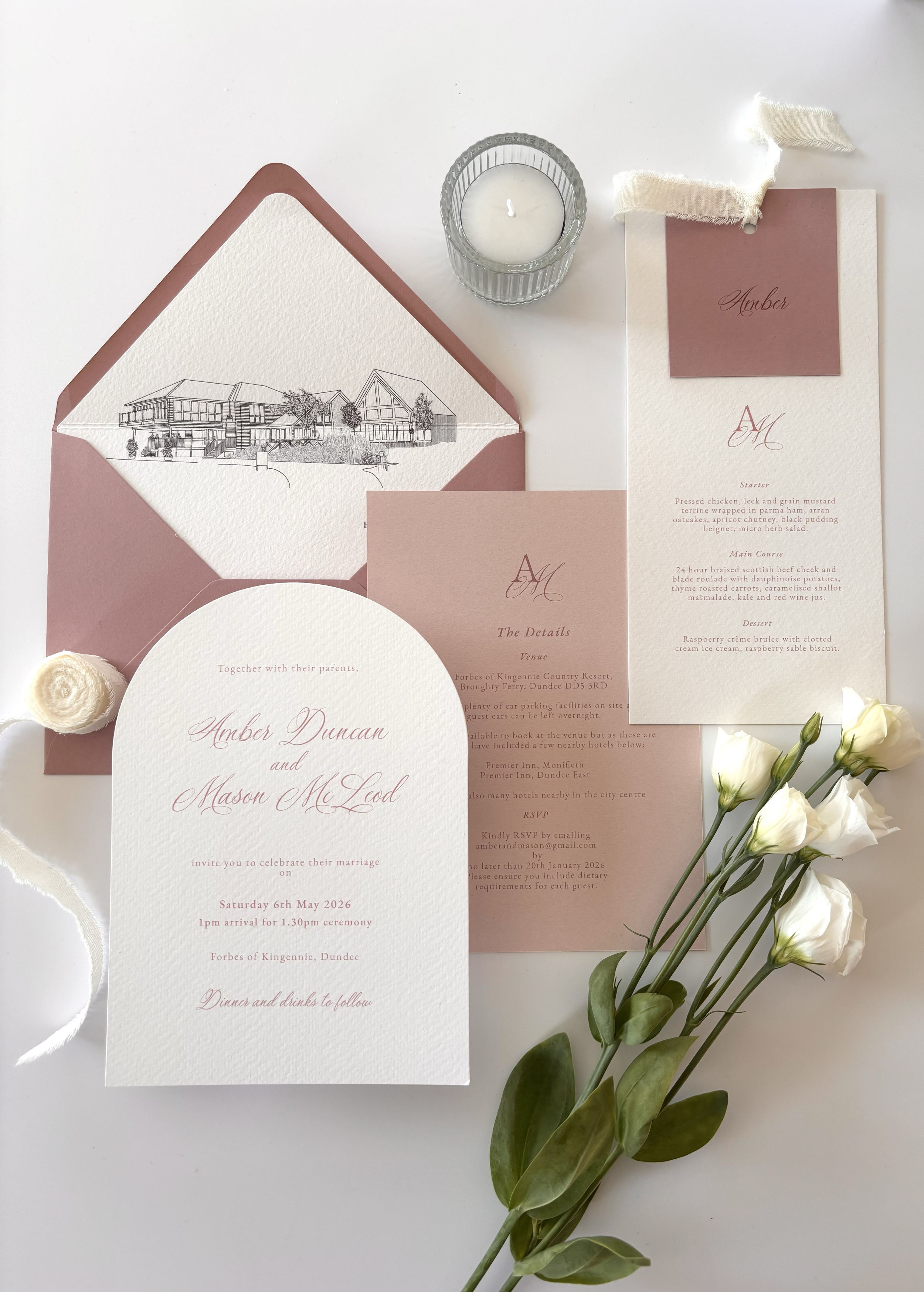 Elegant wedding invitation suite with white textured paper, pink accents, and calligraphy, including an envelope, menu, and RSVP card, accompanied by white flowers and a candle.