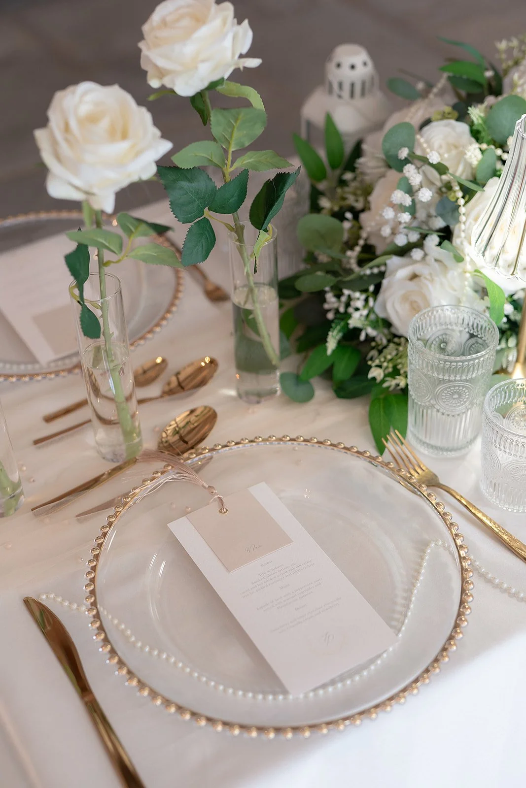 Elegant table setting with a transparent plate bordered with pearls, gold utensils, clear textured glasses, white roses, greenery, and a menu card, suggesting a formal event or wedding reception.