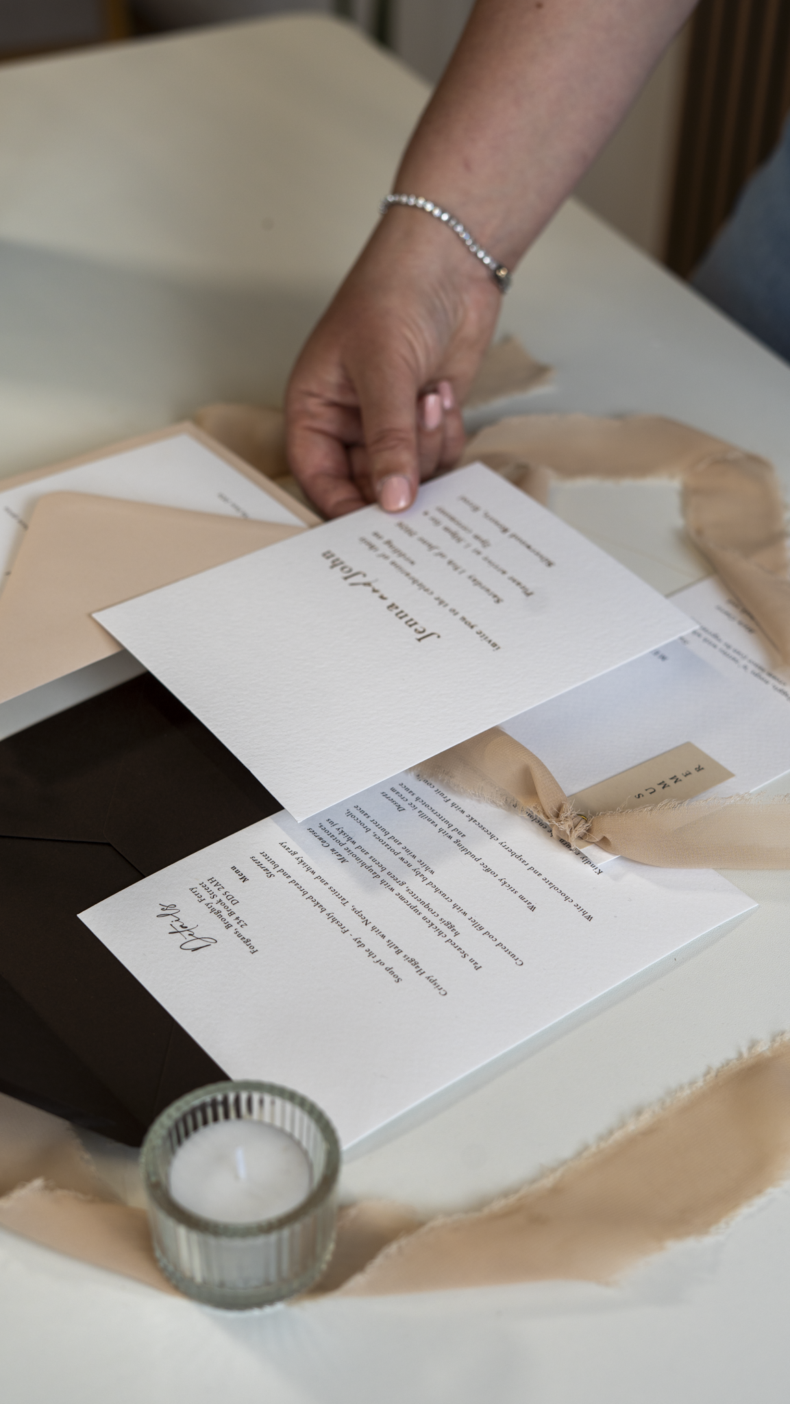 A person's hand adjusting a wedding invitation on a table decorated with a beige cloth, with an unlit candle in a glass holder nearby.