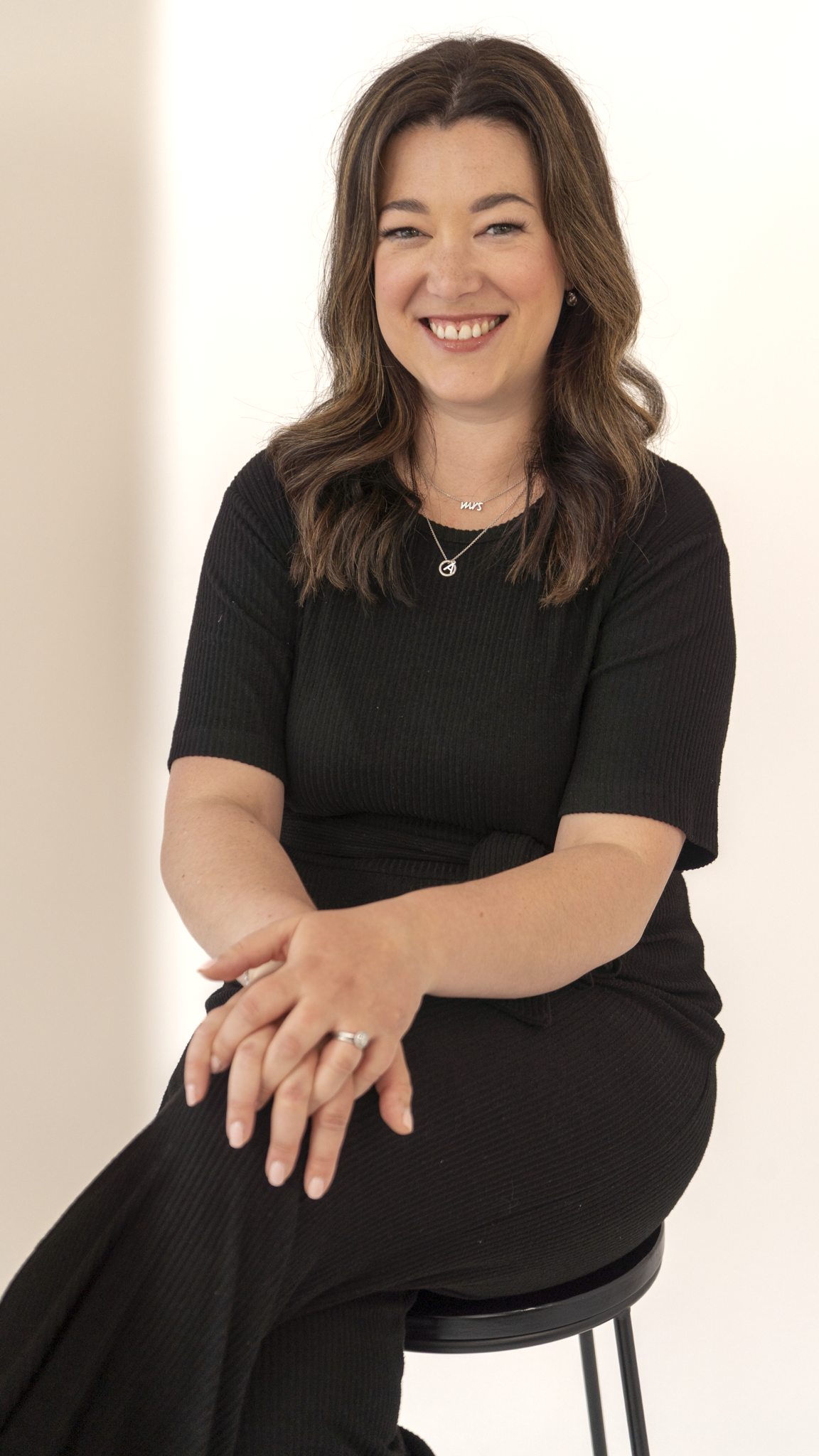 A woman with wavy brown hair, wearing a black short-sleeved top and necklaces, sitting on a black stool with a white background, smiling at the camera.