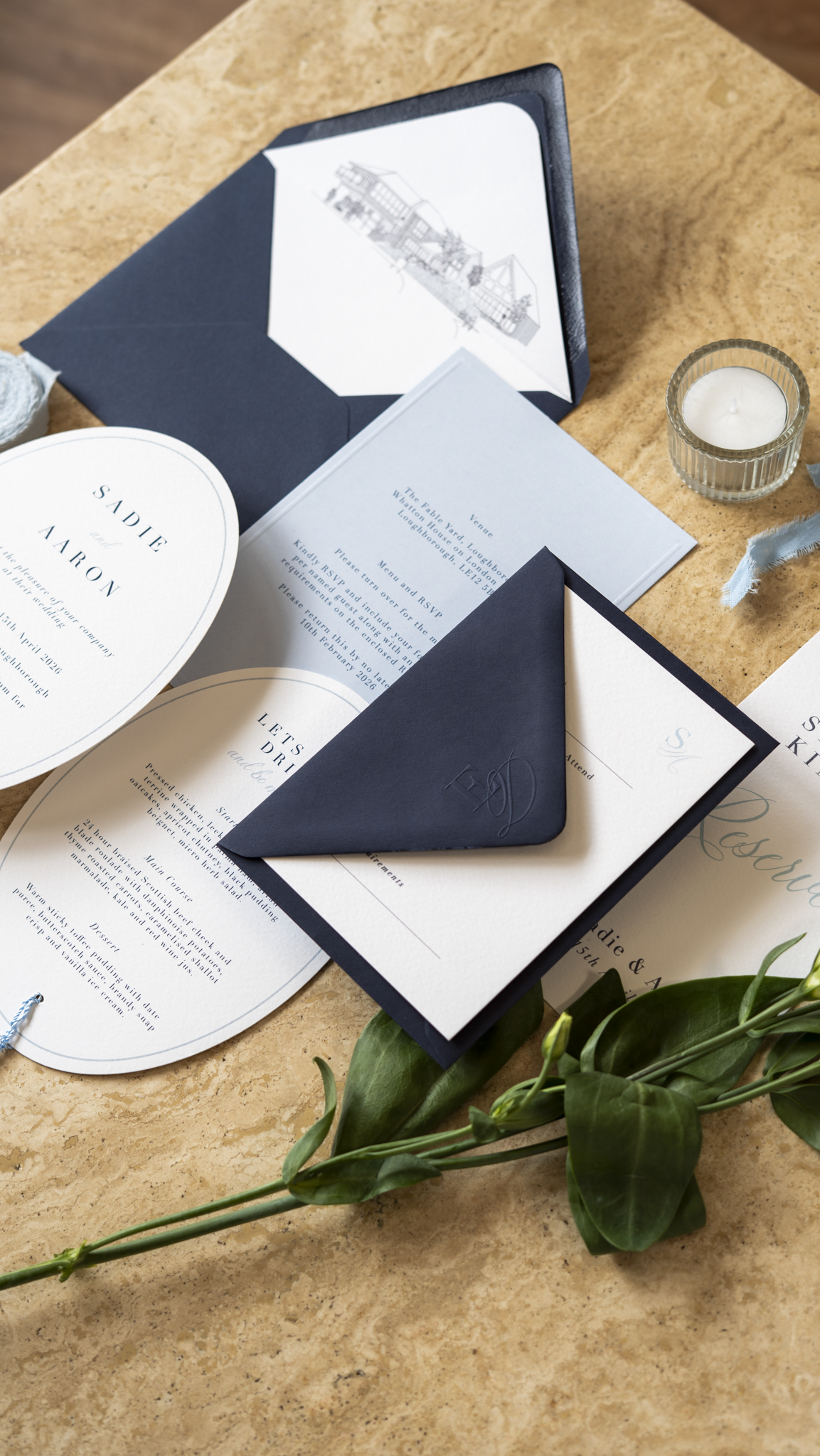 Wedding invitation suite on a wooden table, including a navy envelope, a menu card, a round invite card with blue text, a dark blue pocket fold with a white card inside, a white candle in a glass holder, and some green foliage. The invitation mention