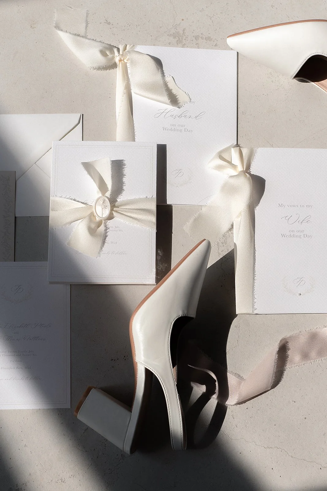 Elegant wedding invitations featuring white paper and satin ribbons, accompanied by white high-heeled shoes, sunlight casting shadows on a neutral surface.
