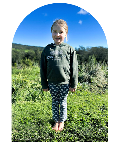 A young girl stands outdoors in a natural environment with green plants and trees in the background, smiling at the camera. She has light brown hair tied up in a ponytail and is wearing a light purple t-shirt and light-colored pants, barefoot on the ground.
