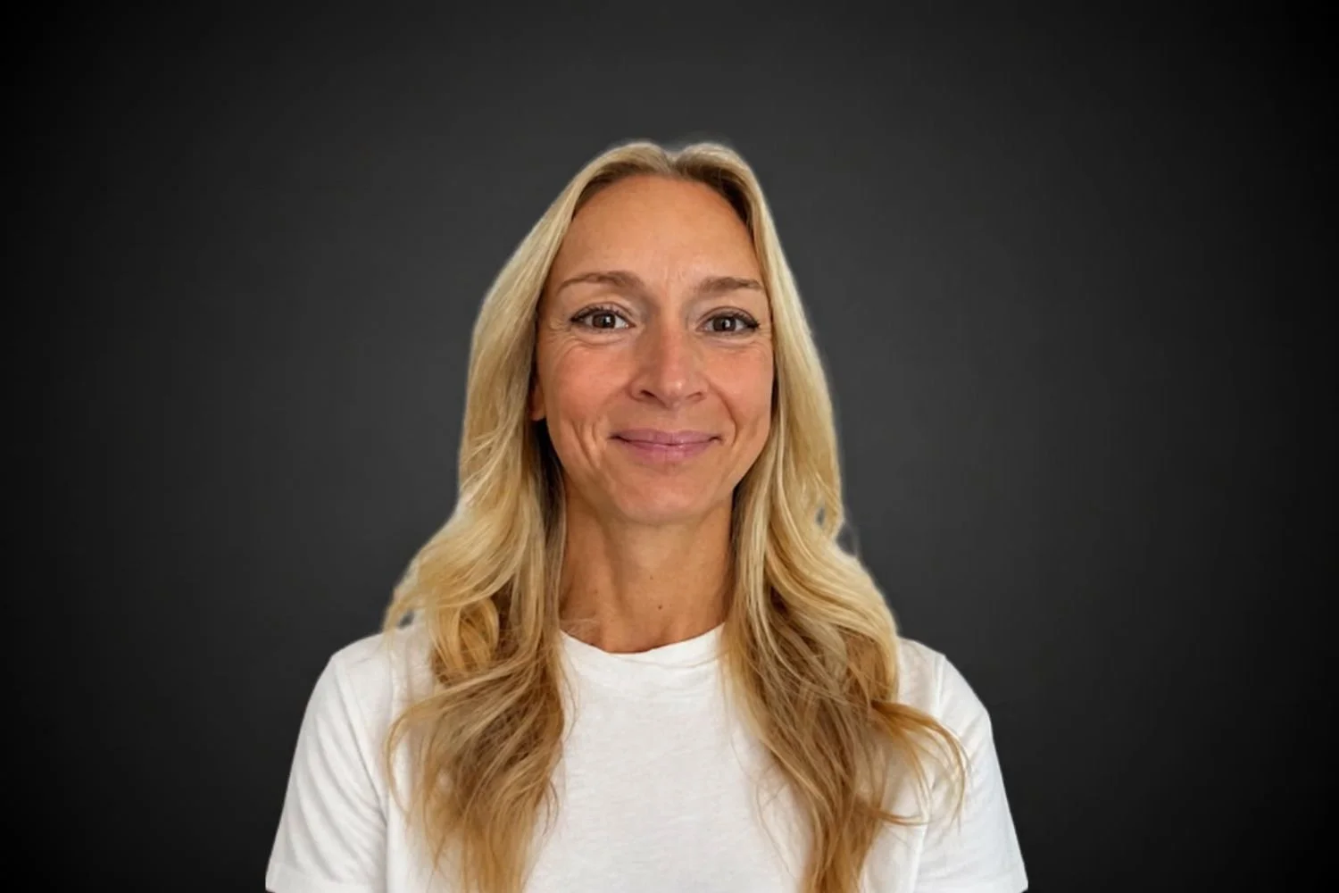 A woman with long blonde hair wearing a white shirt, smiling, against a dark background.