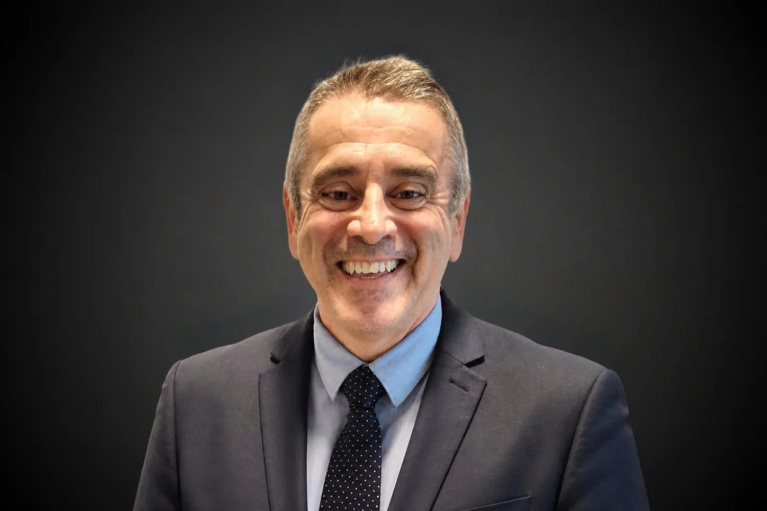 Portrait of a smiling middle-aged man in a gray suit with a light blue shirt and a dark polka dot tie, against a dark background.